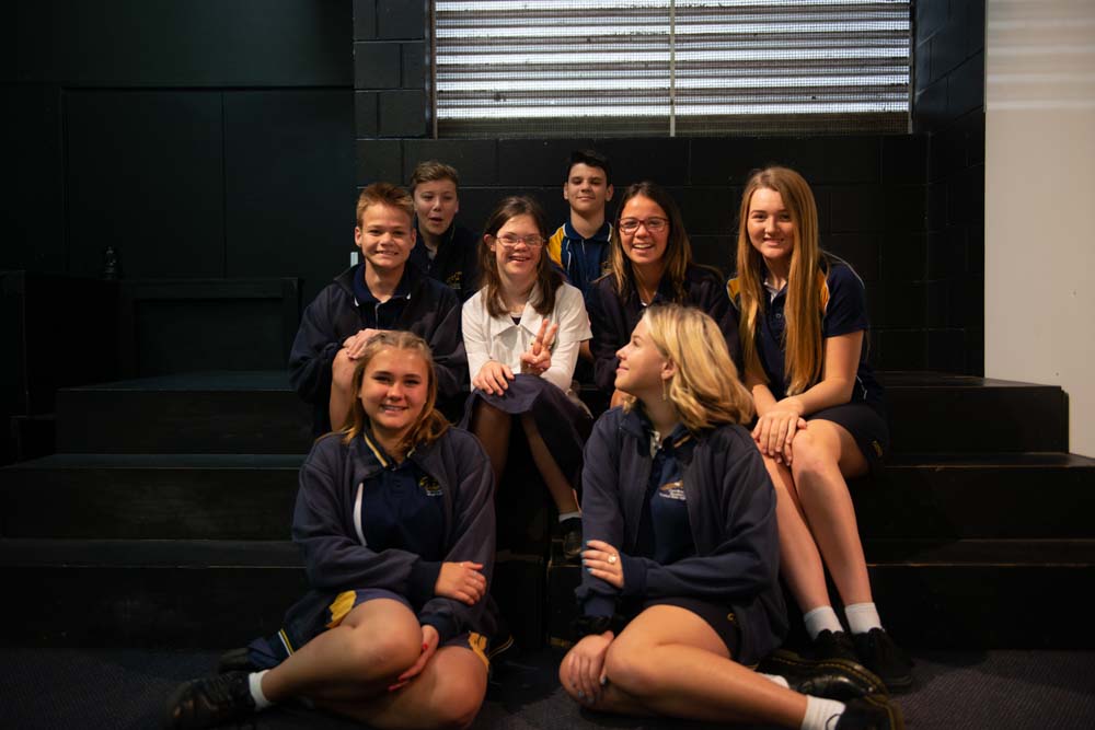  A group of male and female school students sitting on some stairs, all dressed in school uniforms and clearly enjoying each other’s company.  One of the student’s, in the center of the group, has a disability. 