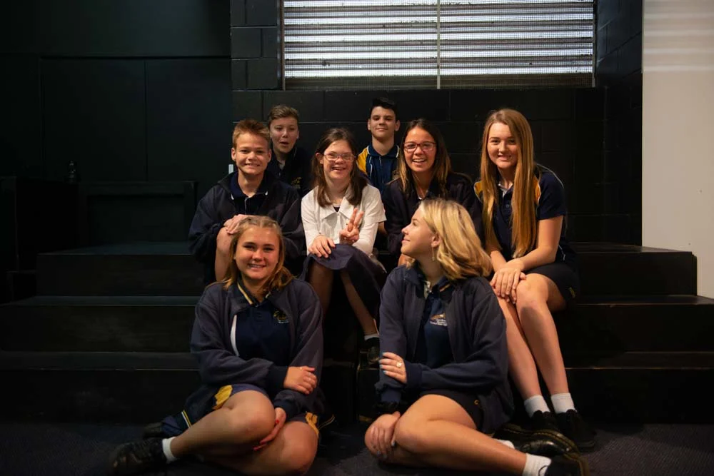 A group of male and female school students sitting on some stairs, all dressed in school uniforms and clearly enjoying each other’s company. One of the student’s, in the center of the group, has a disability.