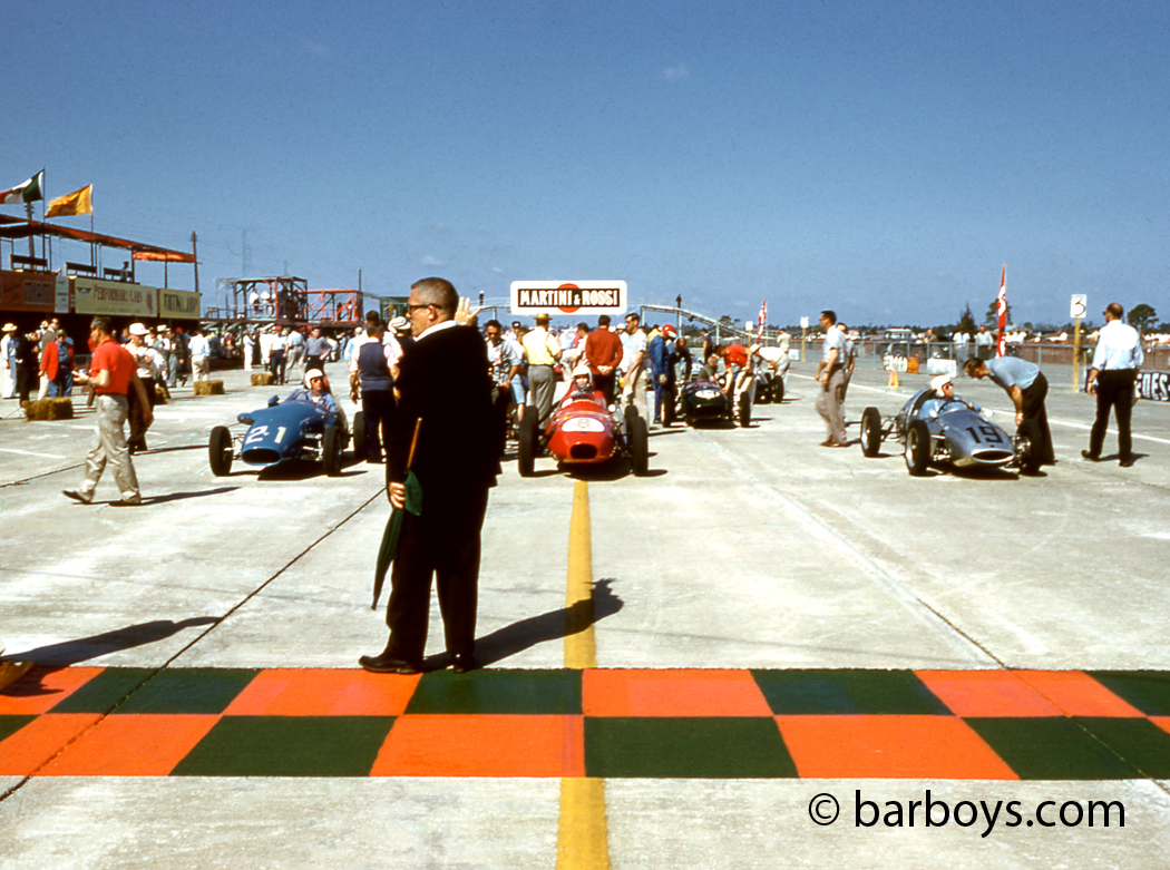 Jesse Coleman stands at the start finish line. The front row has Hansgen in the silver rear engined Cooper, Jim Hall in the red Elva and Ed Crawford in the blue Stanuellini.