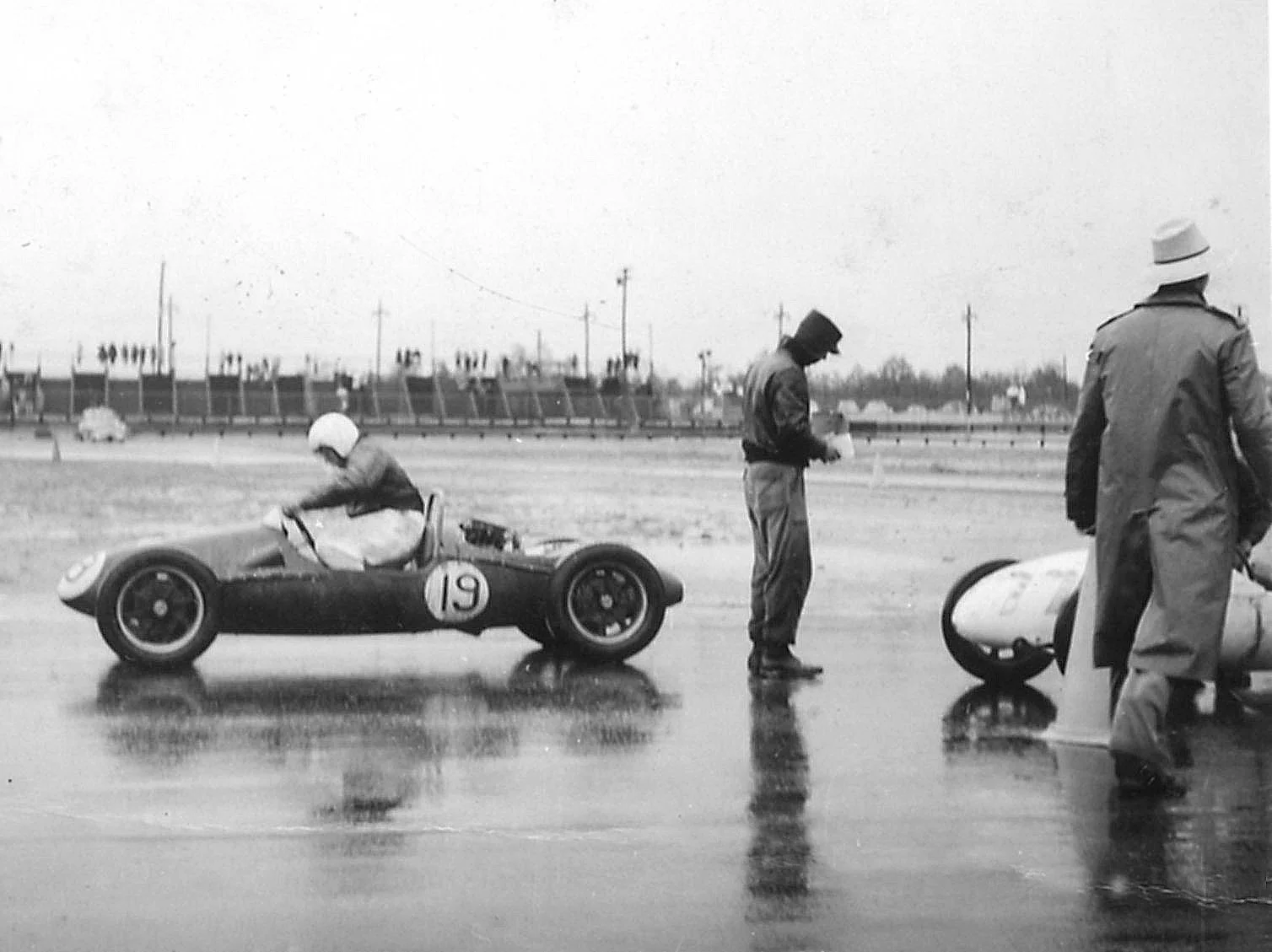 The rain is falling as Jim Haynes gets into his car. He is on the back straight of the oval. You can see the grandstands in the background.