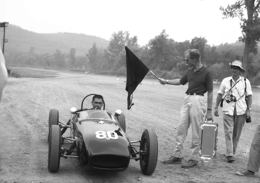 Harry Carter arrives back at the pits in his Lotus 18, his sponsor Freddy Bowles loaded down with cameras.