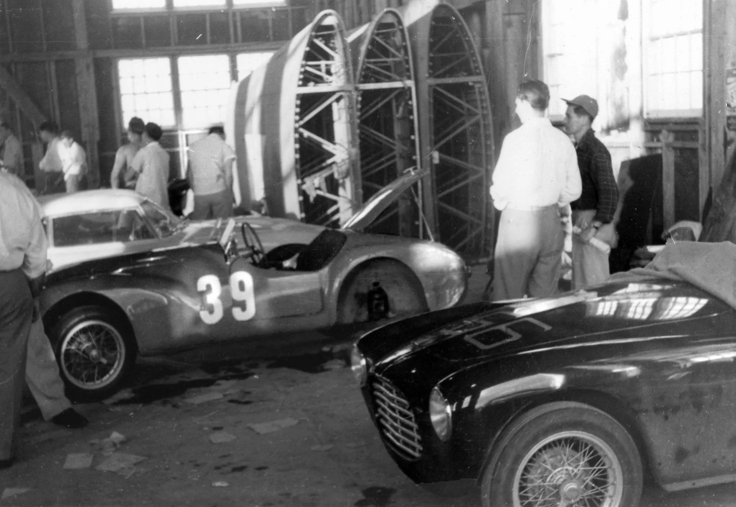  1953: Tom Scatchard &amp; Henry Wessells #56 Siata and Otto Linton and John Edgar’s #39 Siata Amica special sit in a hanger where you can see 3 airplane wing pieces stored behind.  The air base was now the Sebring regional airport and remained activ