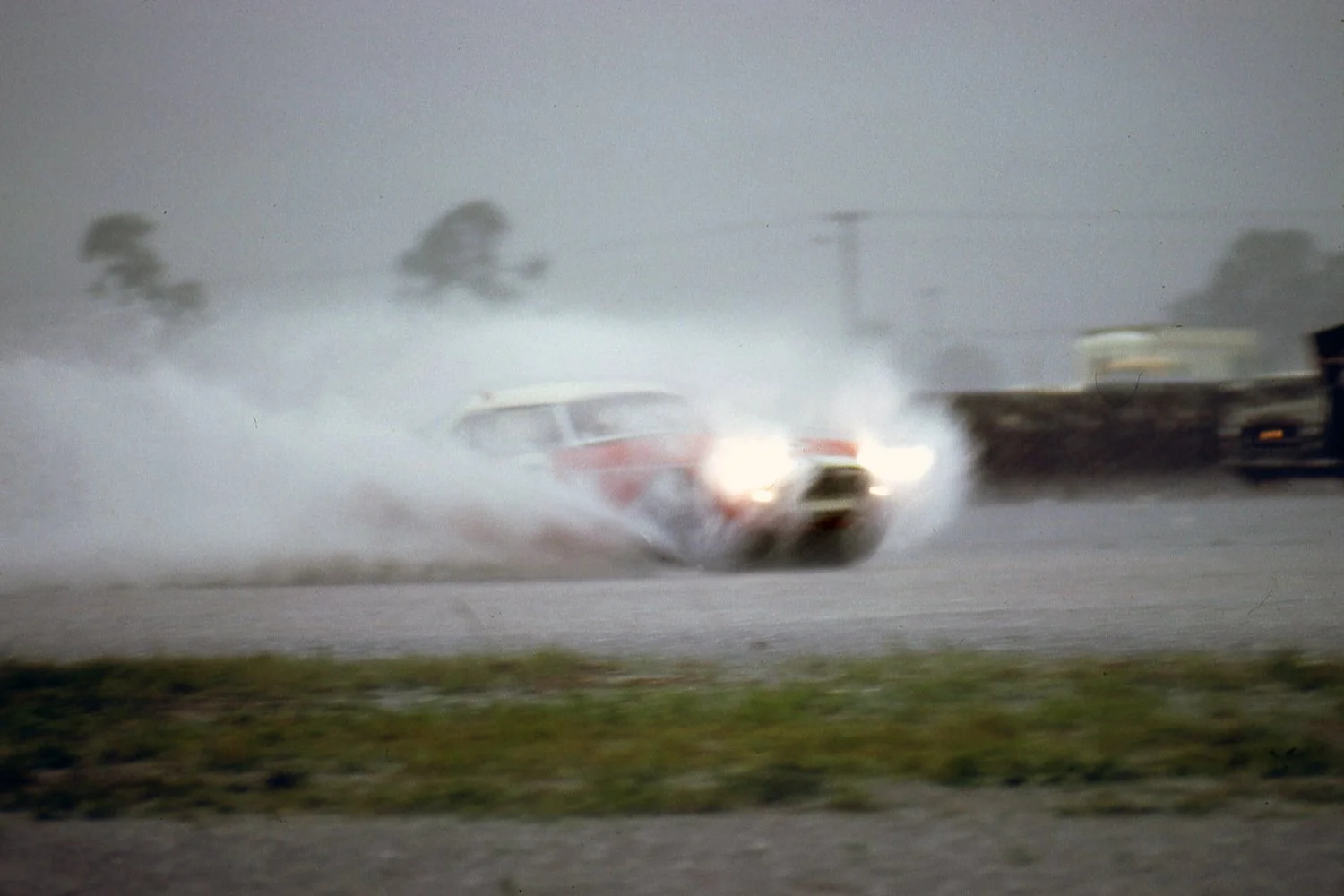  1965:  The track was flooded but unlike today, the race carried on.  And it created the possibility of some great photos.  A factory MGB looks more like a speedboat as it runs with lights blazing. 
