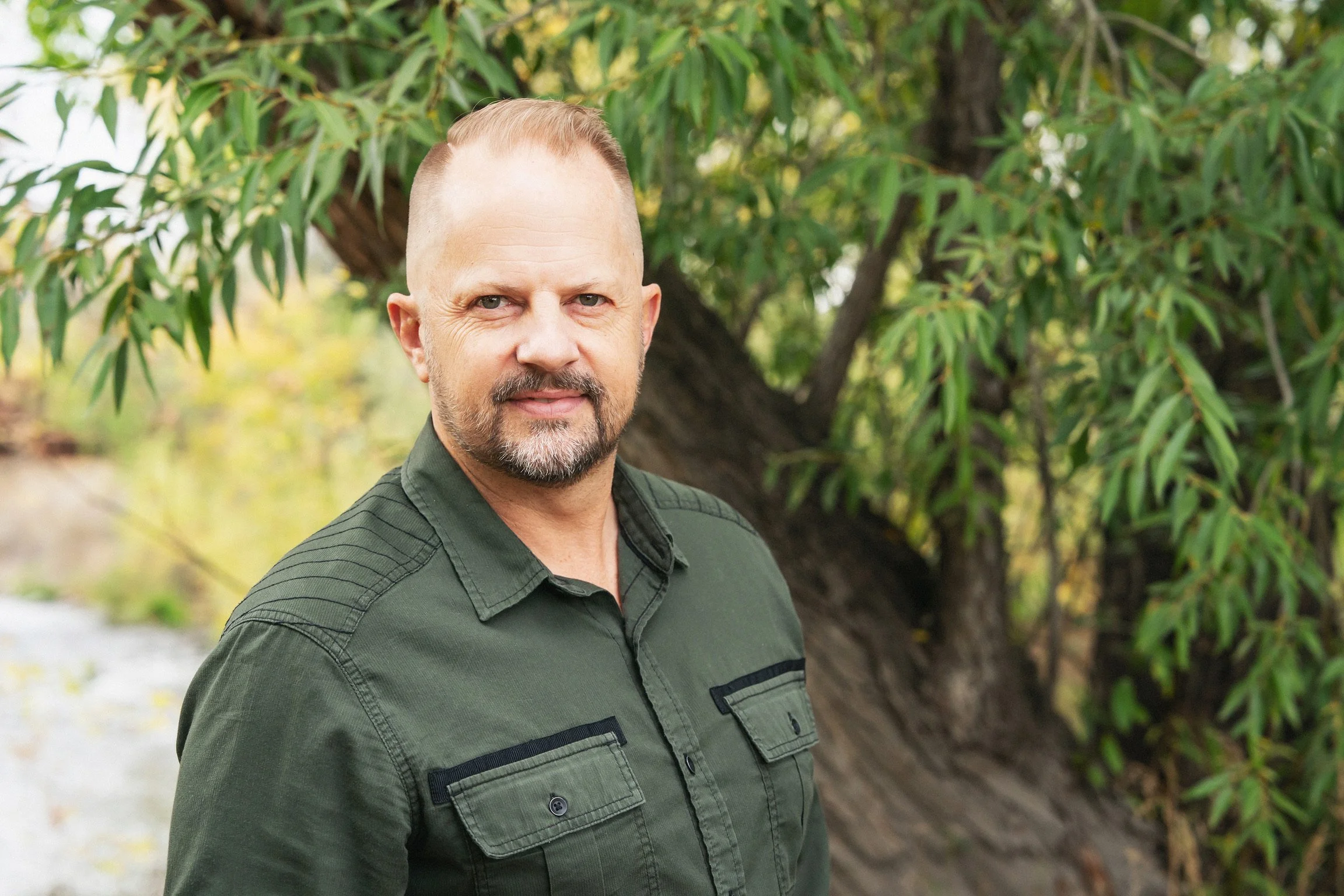 A middle-aged man with short blond hair, a beard, and blue eyes wearing a green button-up shirt standing outdoors in front of a large leafy tree.