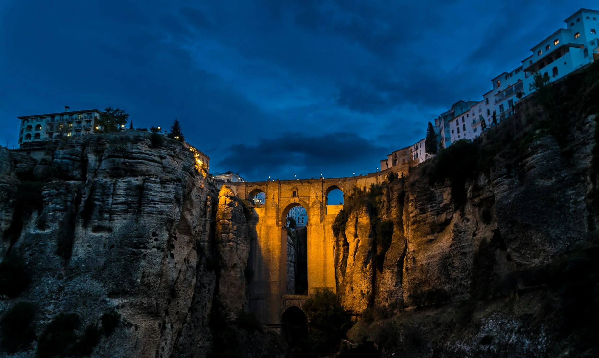 Night view of a gorge with a bridge illuminated in golden light, surrounded by houses and trees on rocky cliffs, under a dark blue cloudy sky.