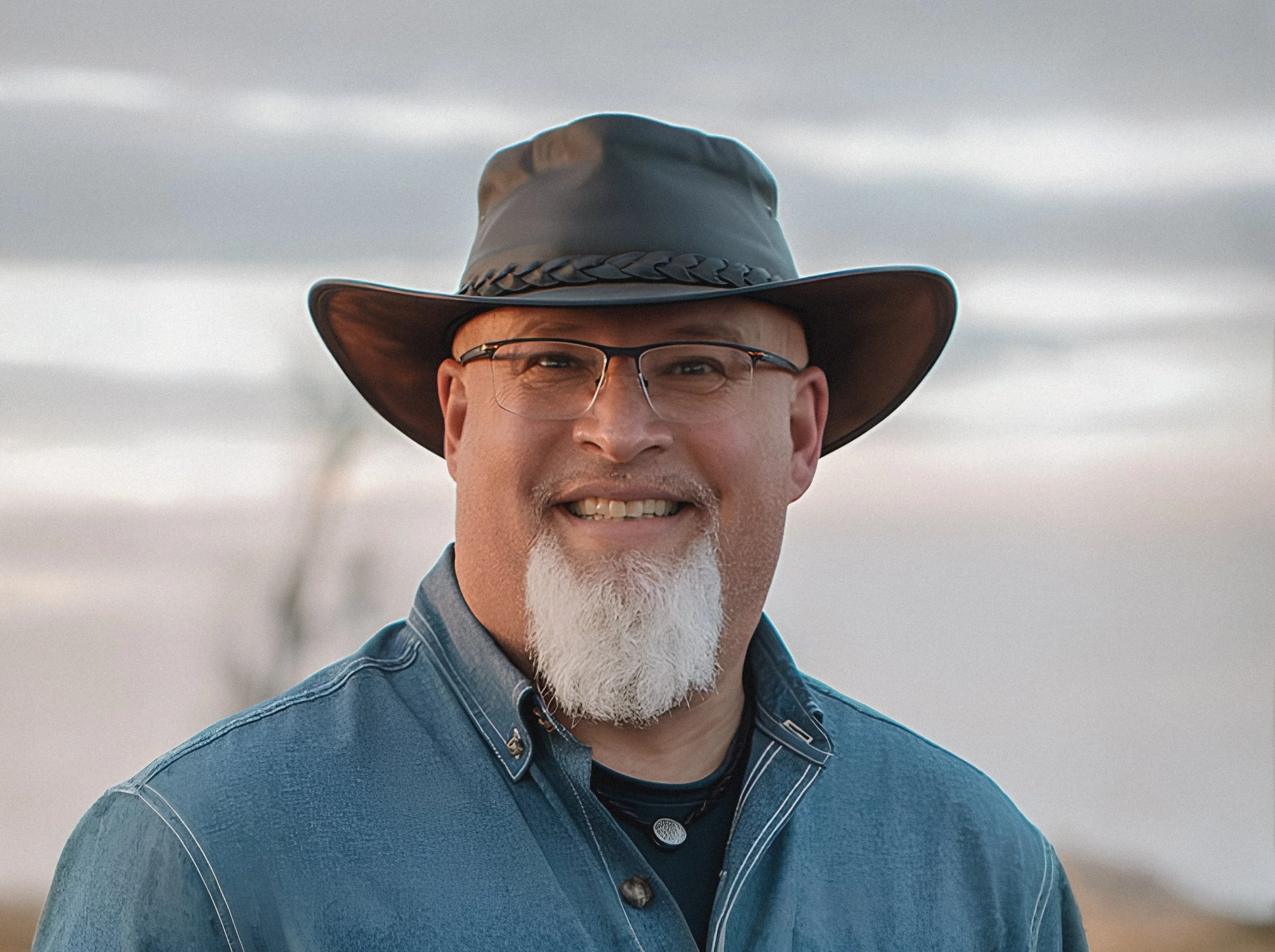 A man wearing a wide-brimmed hat, glasses, a denim shirt, and a black t-shirt, outdoors with a body of water and cloudy sky in the background.