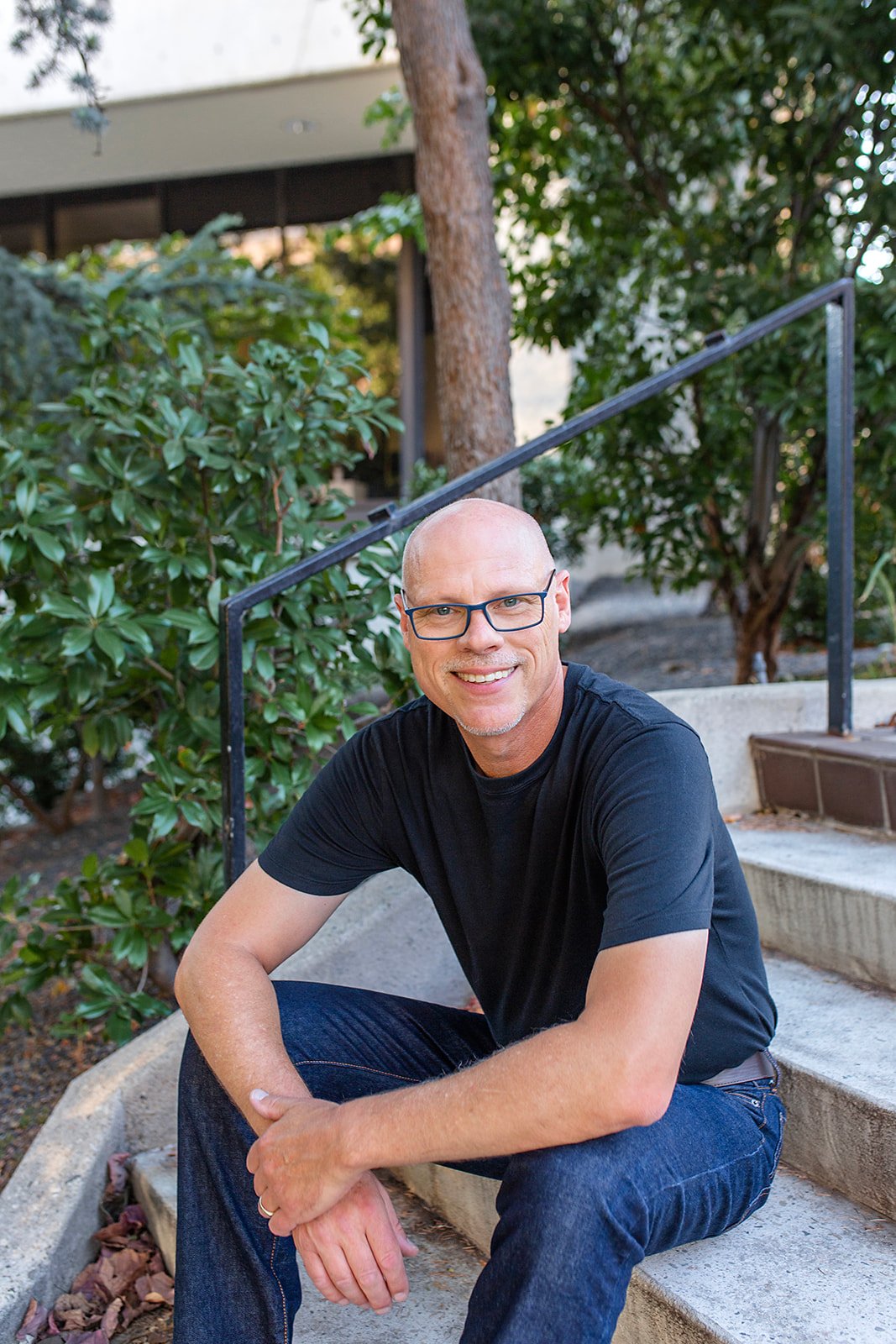 A man with glasses and a black t-shirt sitting on outdoor stairs surrounded by green bushes and trees.