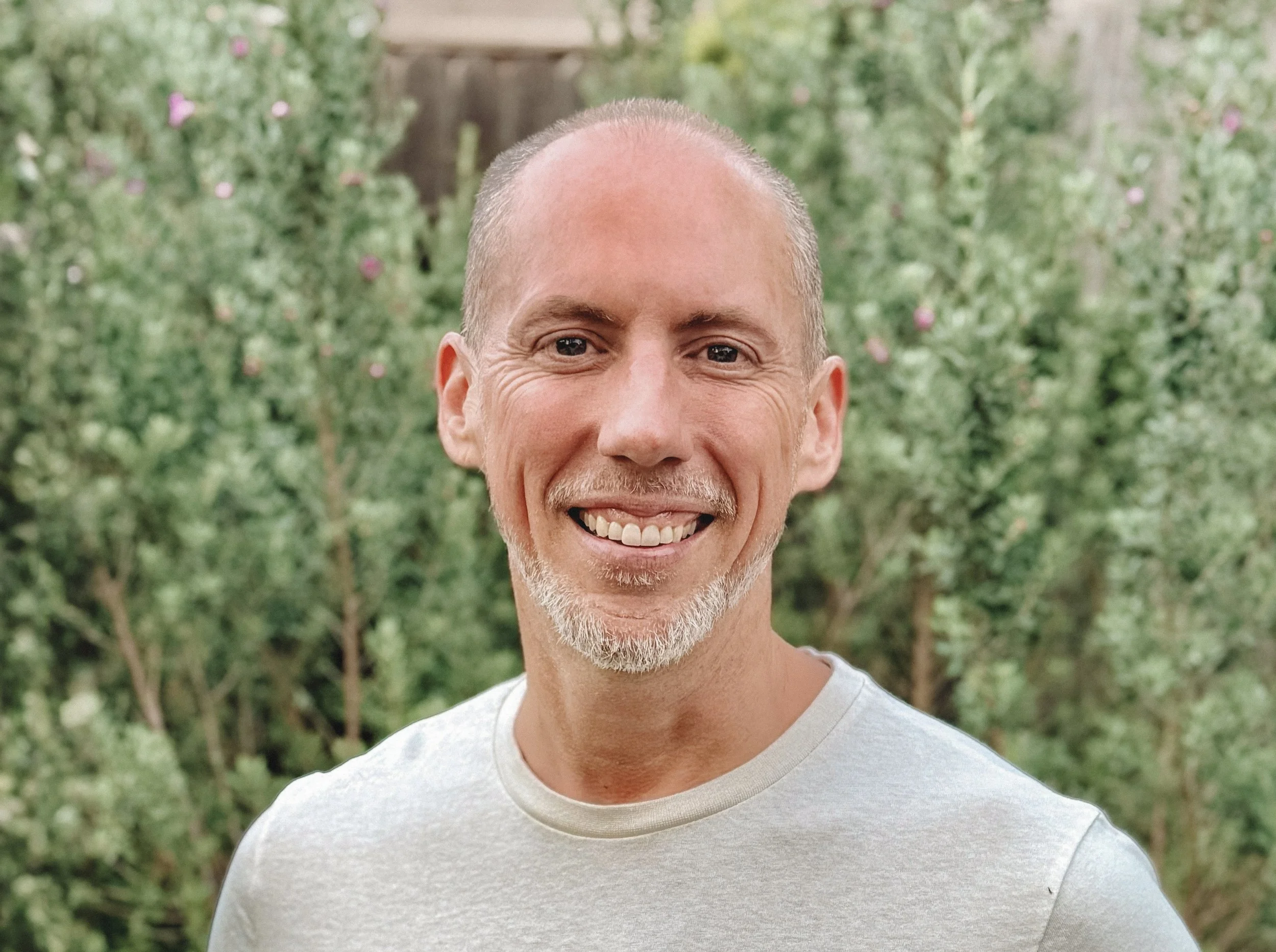 Close-up of smiling middle-aged man with short hair and a beard, wearing a white t-shirt, standing outdoors with green trees in the background.