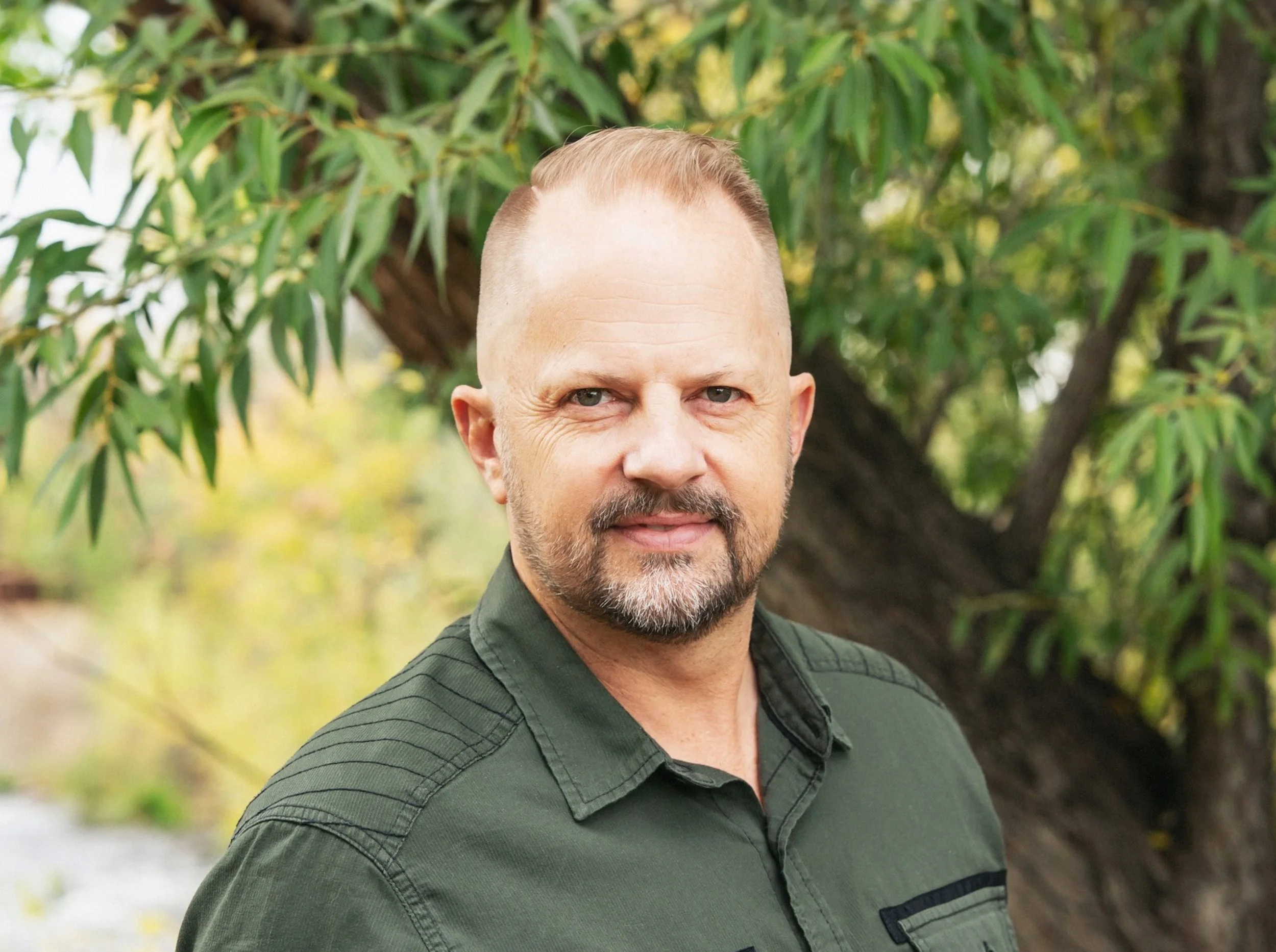 A middle-aged man with short blond hair, a beard, and blue eyes wearing a green button-up shirt standing outdoors in front of a large leafy tree.