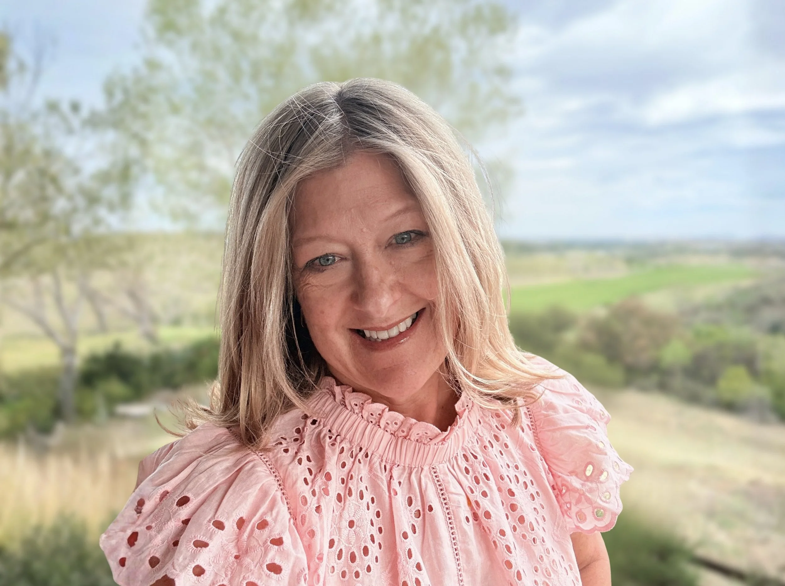 A smiling woman with long blond hair and blue eyes, wearing a pink eyelet blouse, standing outdoors with a blurred background of trees and a blue sky.