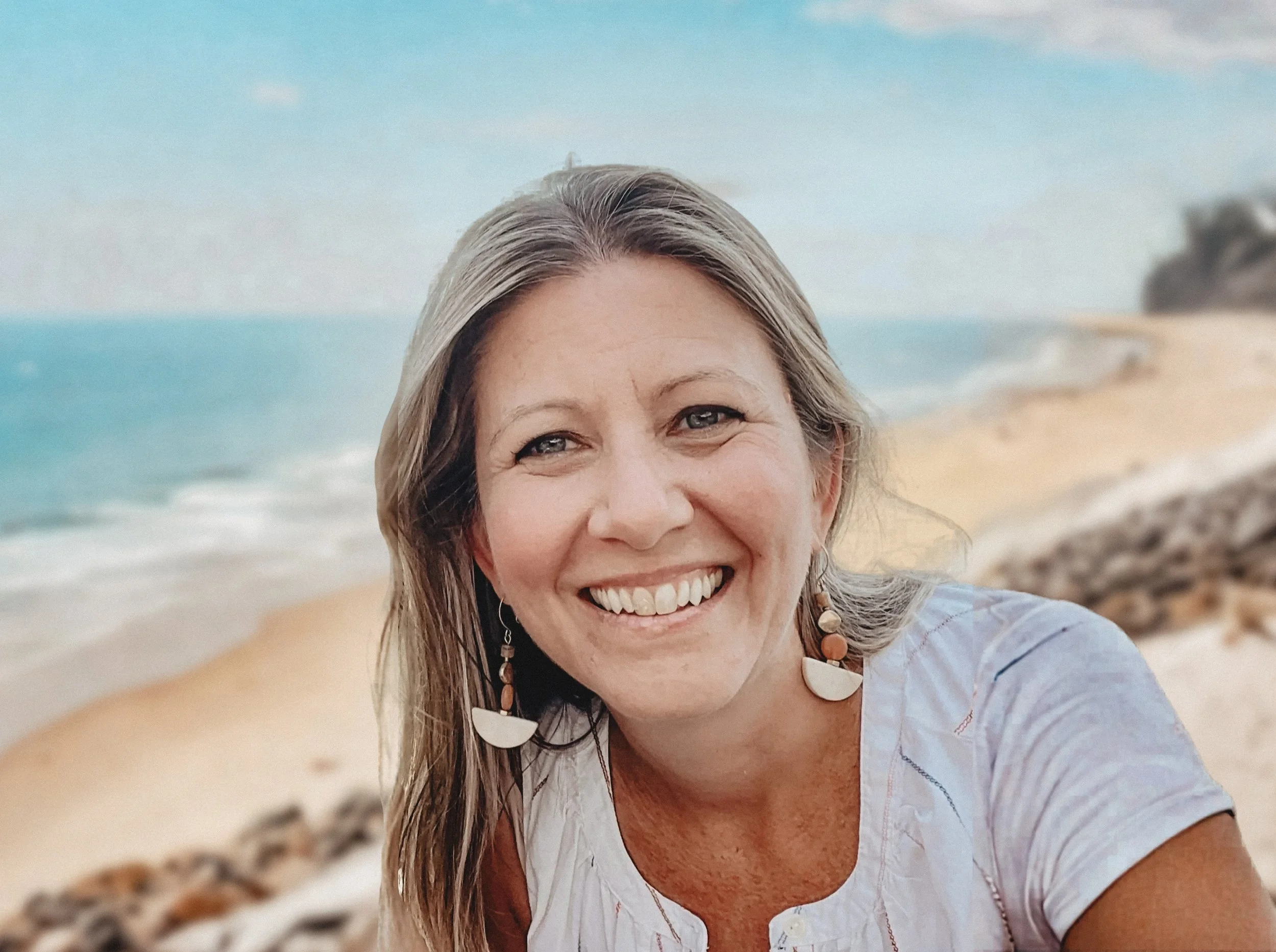 Smiling woman with long hair and earrings at the beach, with ocean waves and rocks in the background.