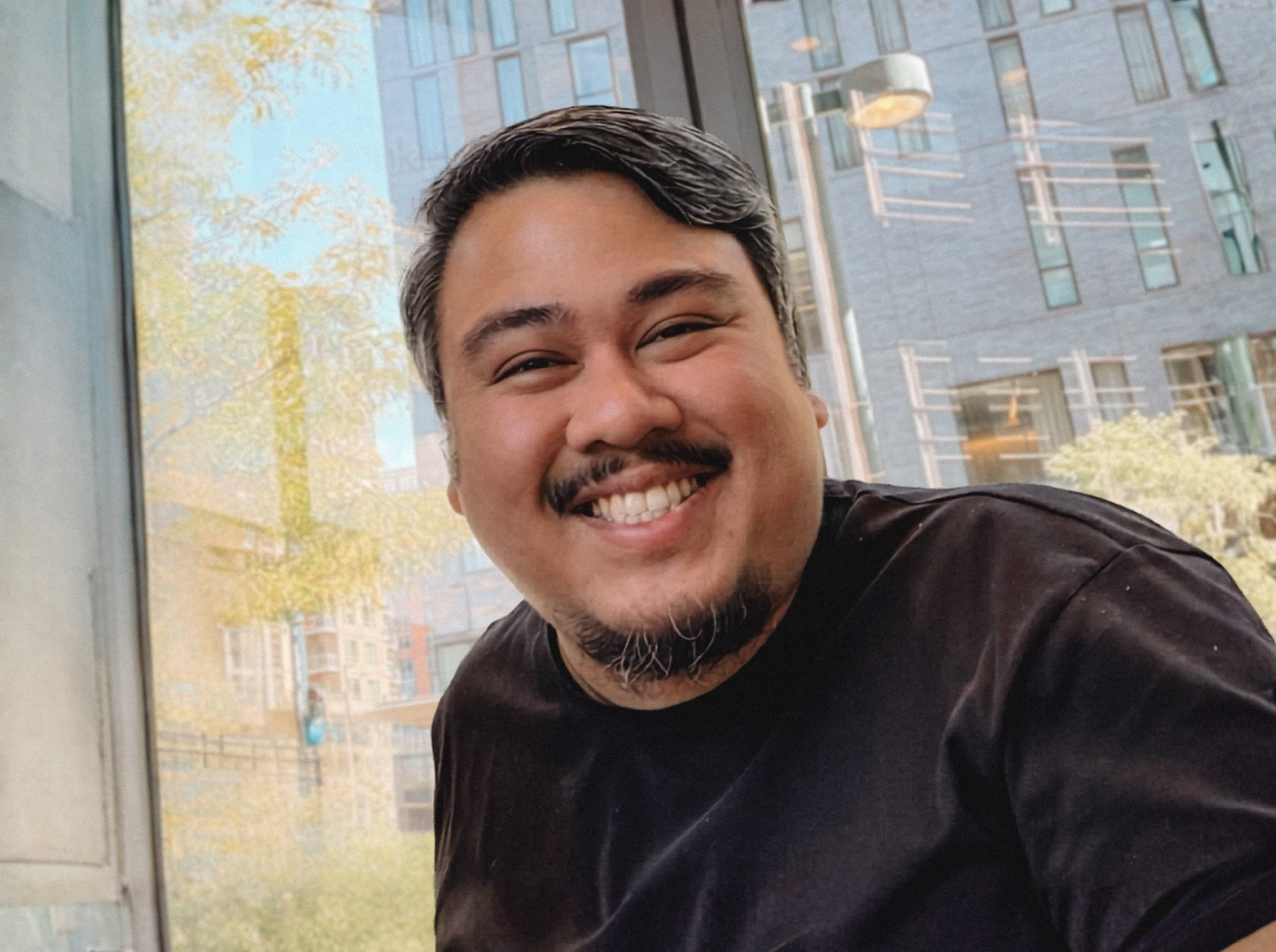Close-up of a smiling man with dark hair and a beard, sitting indoors with large windows behind him showing a cityscape with a building and trees.