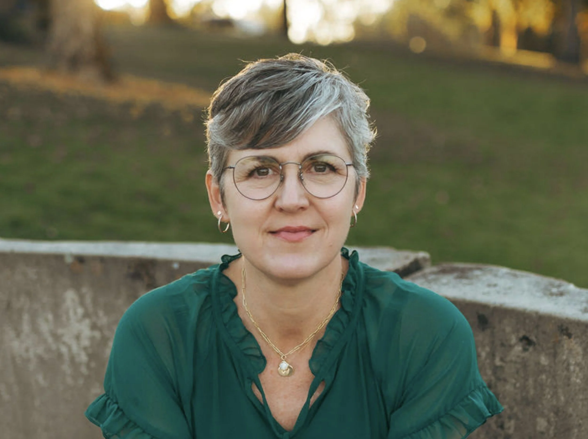 A woman with short gray hair, wearing glasses, a green blouse, and beige pants, sitting on a stone bench in a park during sunset.