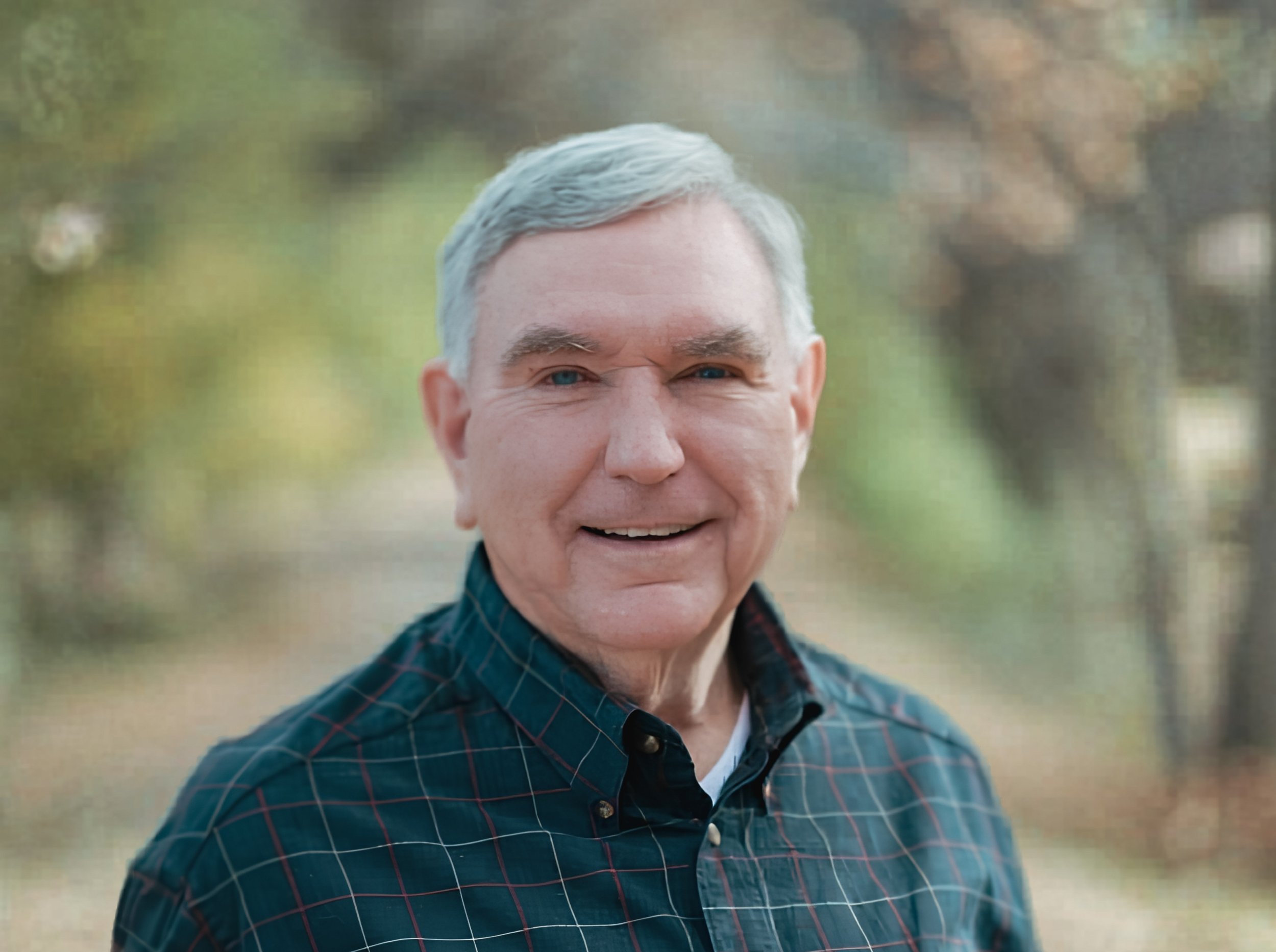Close-up of a smiling older man with gray hair, wearing a dark blue plaid shirt, outdoors with blurred green trees in the background.