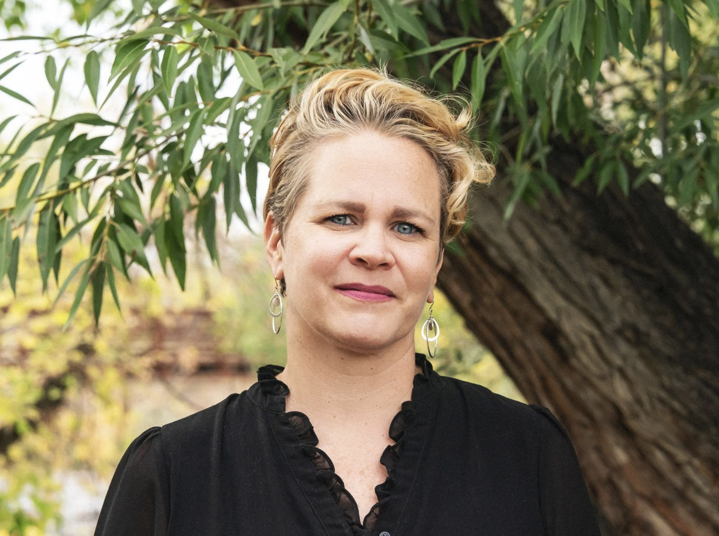 A woman with short blonde hair and blue eyes standing outdoors near a tree with green leaves.
