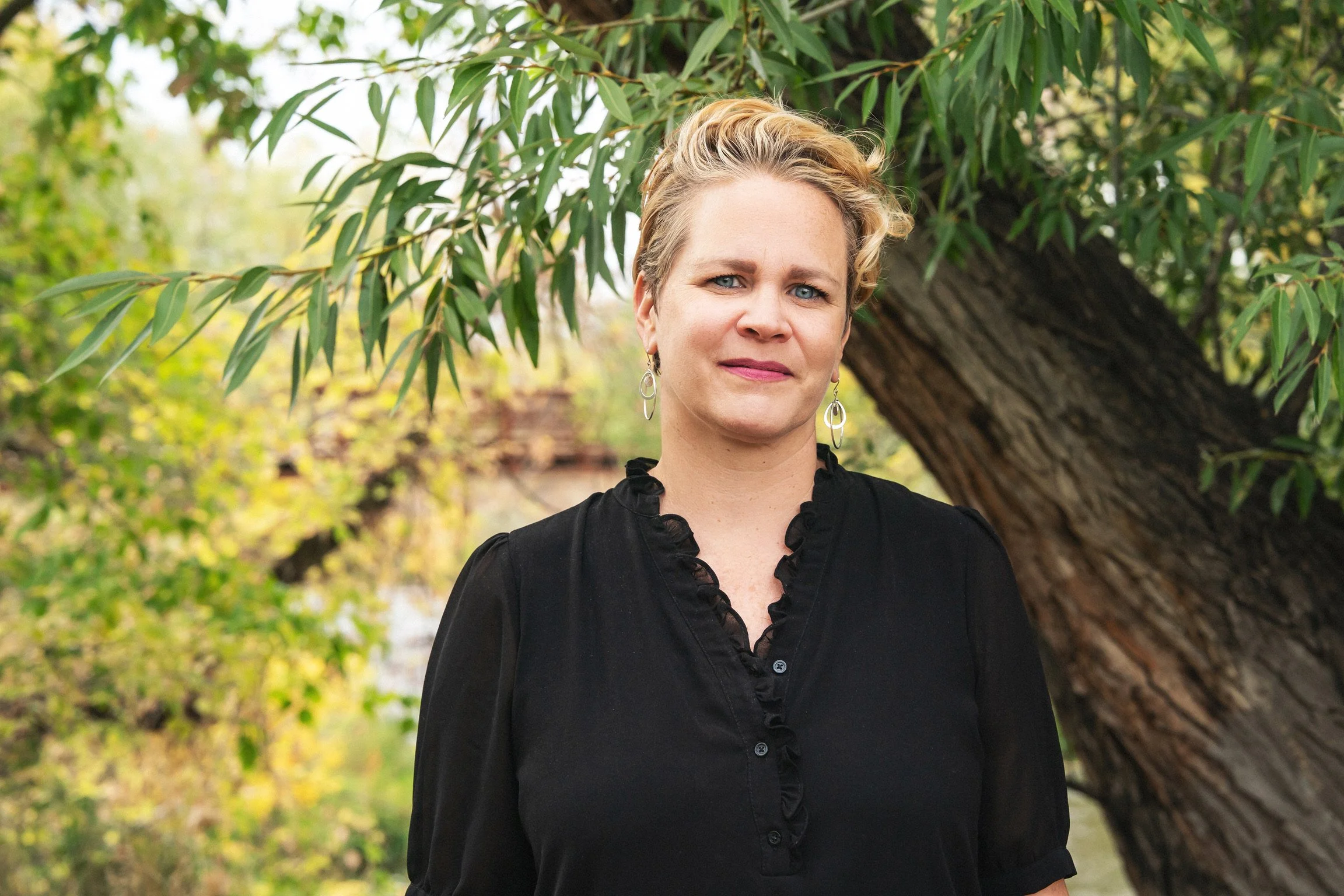 A woman with short blonde hair and blue eyes standing outdoors near a tree with green leaves.