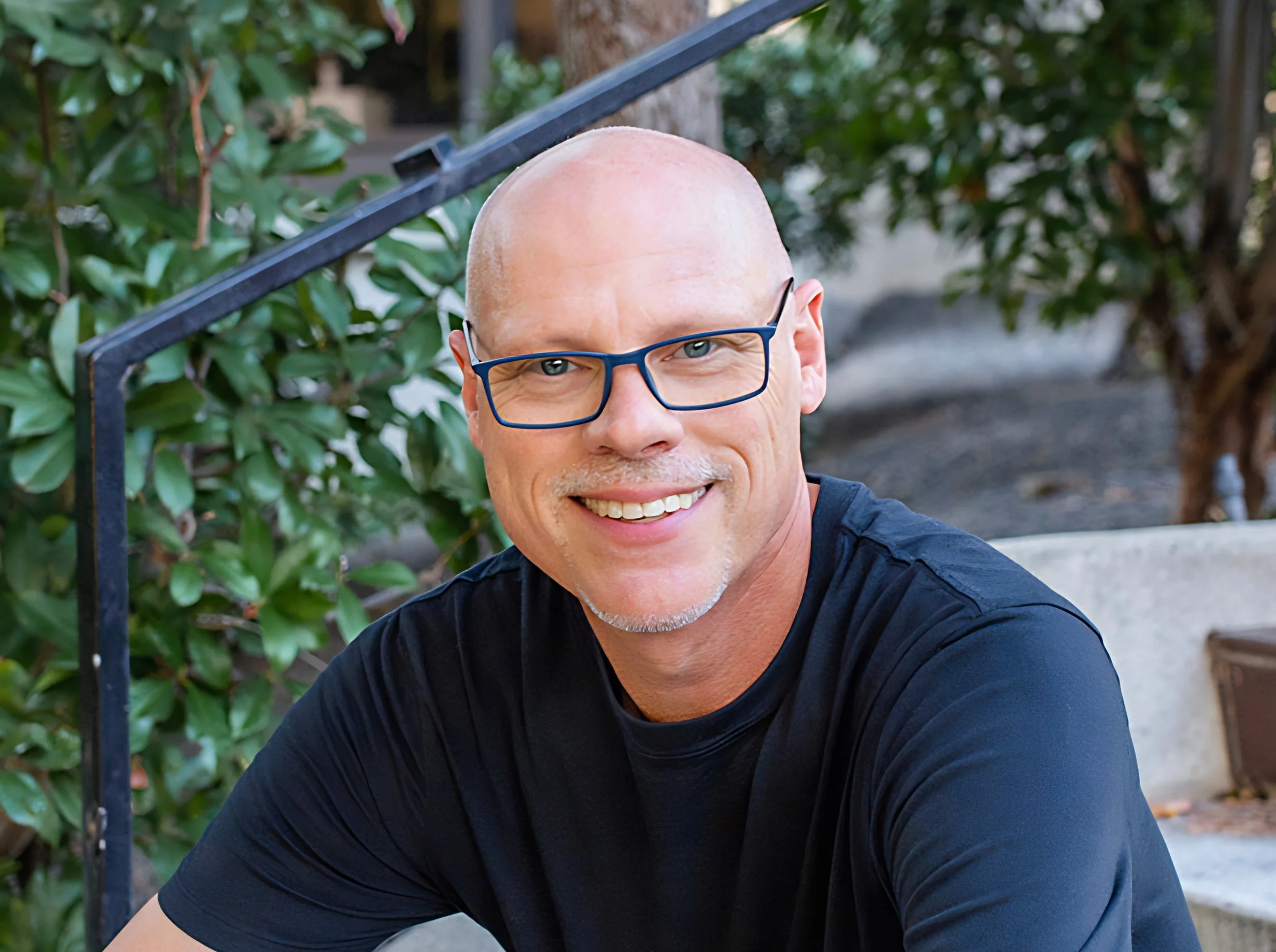 A man with glasses and a black t-shirt sitting on outdoor stairs surrounded by green bushes and trees.