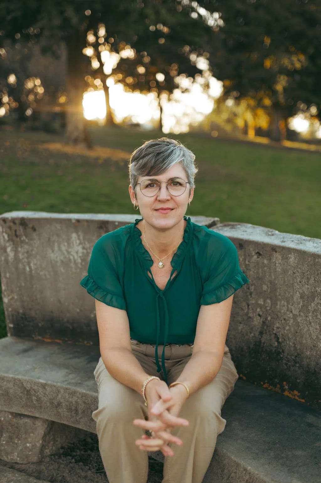 A woman with short gray hair, wearing glasses, a green blouse, and beige pants, sitting on a stone bench in a park during sunset.