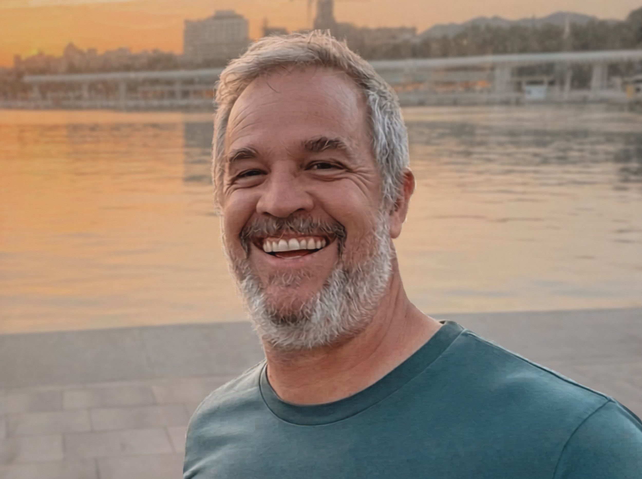 Smiling middle-aged man with gray hair and beard taking a selfie by the water at sunset, city skyline and mountains in the background.