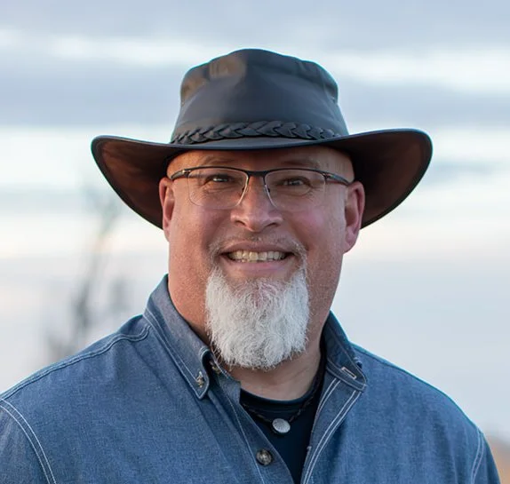 A man wearing a wide-brimmed hat, glasses, a denim shirt, and a black t-shirt, outdoors with a body of water and cloudy sky in the background.