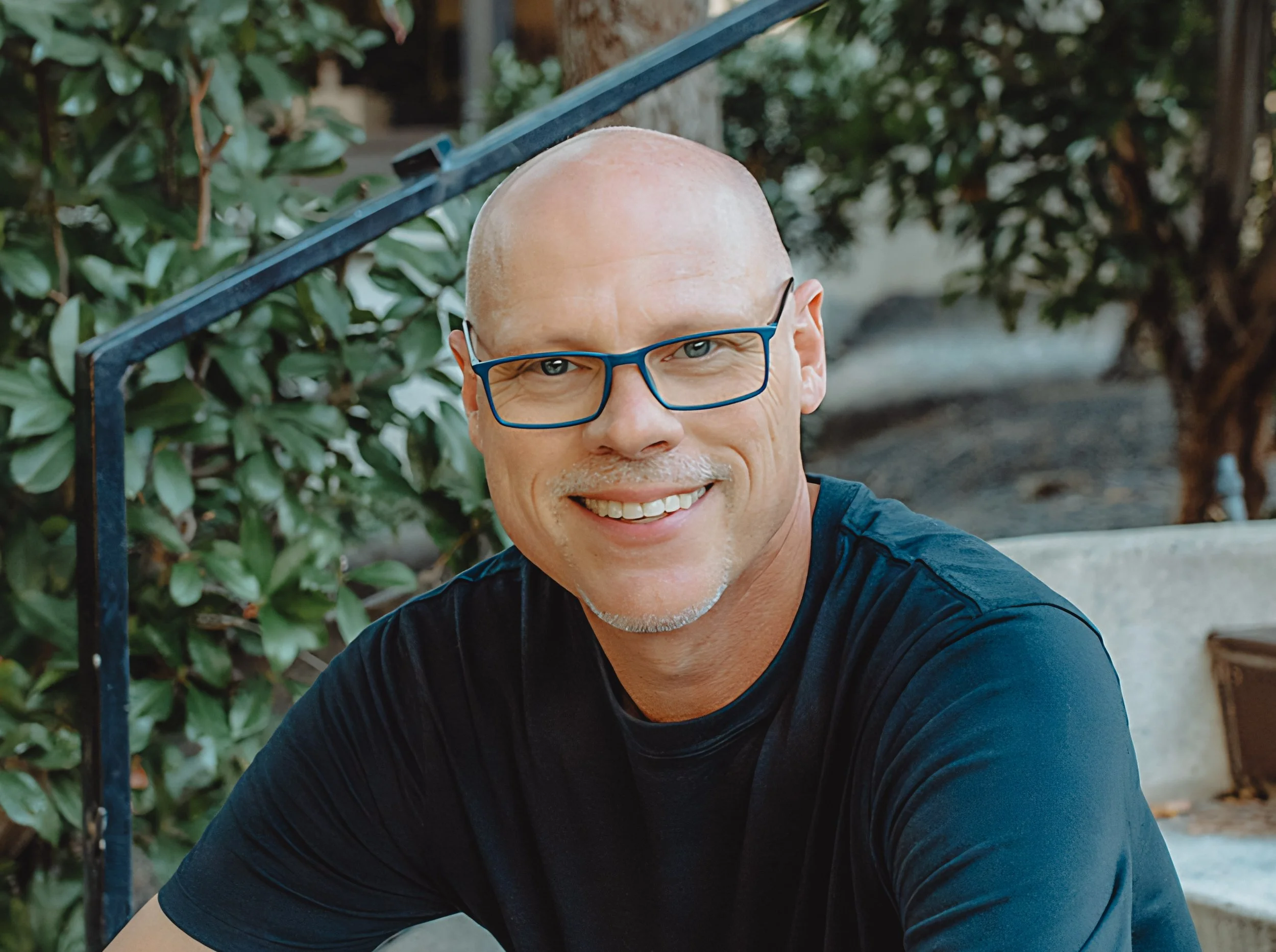 A man with glasses and a black t-shirt sitting on outdoor stairs surrounded by green bushes and trees.