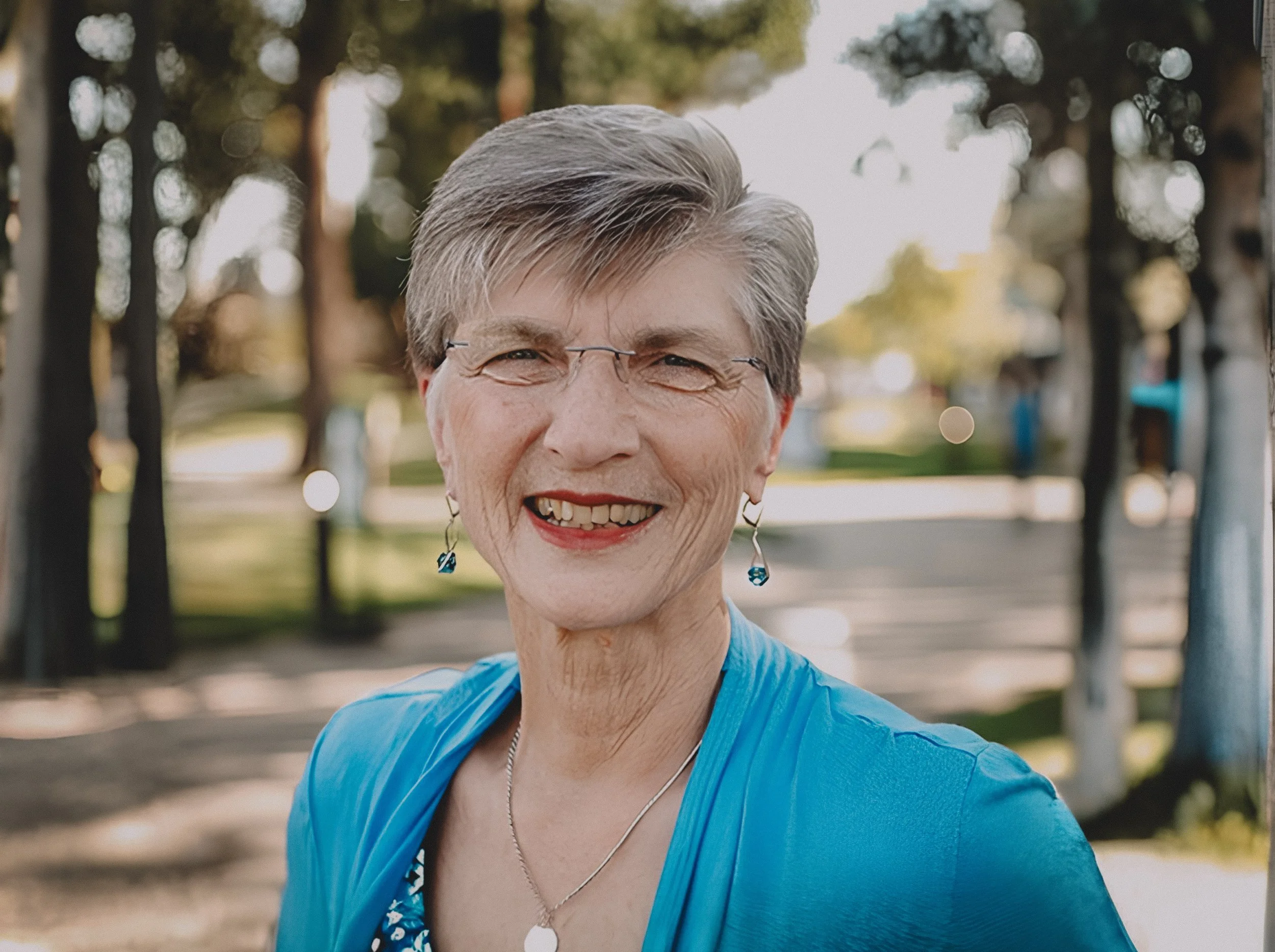 Close-up of a smiling elderly woman with short gray hair, wearing glasses, earrings, a necklace, and a blue athletic top outdoors with blurred trees and a fence in the background.