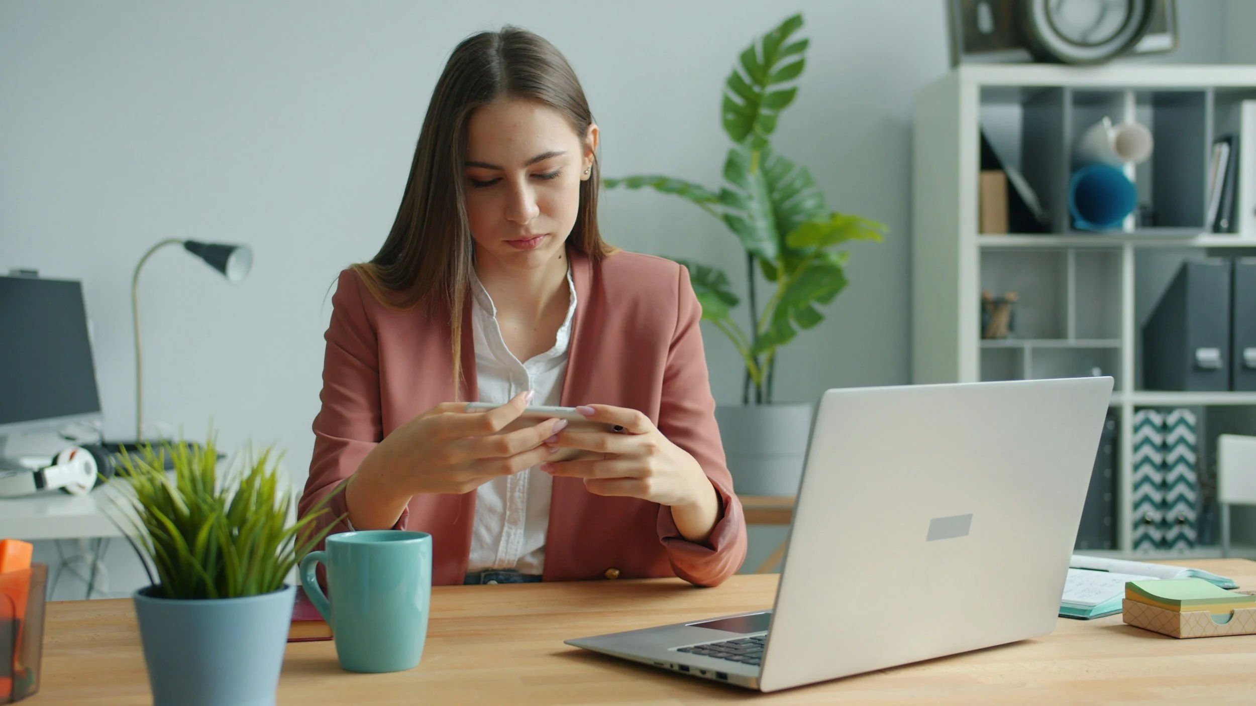 A woman in a home office looking at her cellphone instead of her computer monitor, illustrating a procrastination habit.