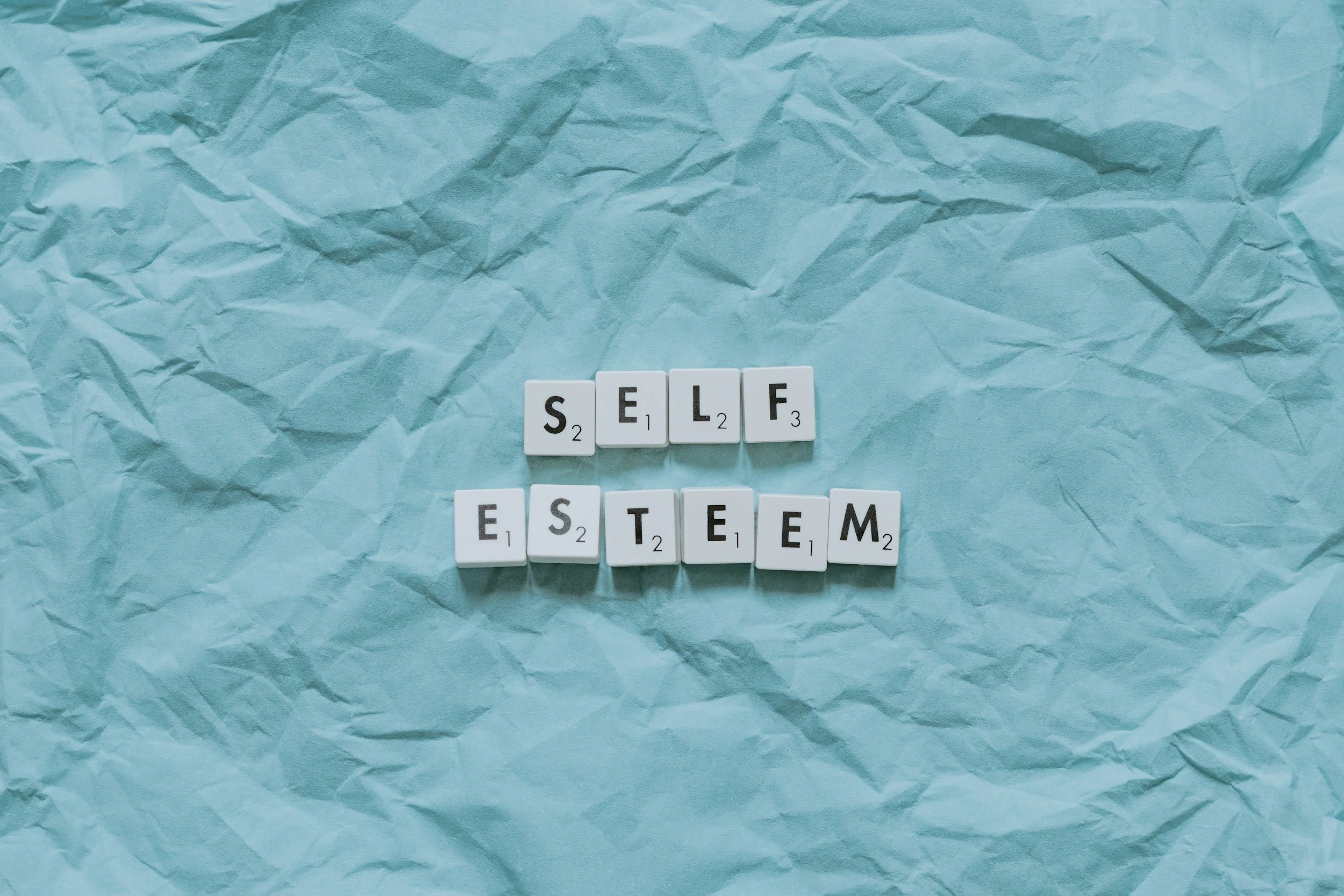 A top-down view featuring wooden Scrabble tiles arranged in two stacked rows to spell "SELF ESTEEM" across the center of a textured, light blue crinkled paper background.