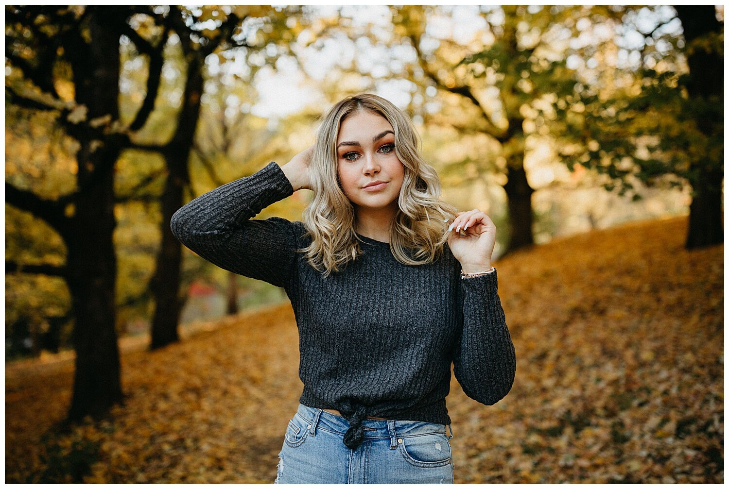  Girl wearing a grey sweater surrounded by orange leaves for her senior photos. 