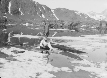 An Inuit man squats on the ice next to a kayak. He dangles a fishing line into the water. Watkins stands behind by another kayak also fishing with a line. Collection- British Arctic Air Route Expedition 1930-31.jpeg