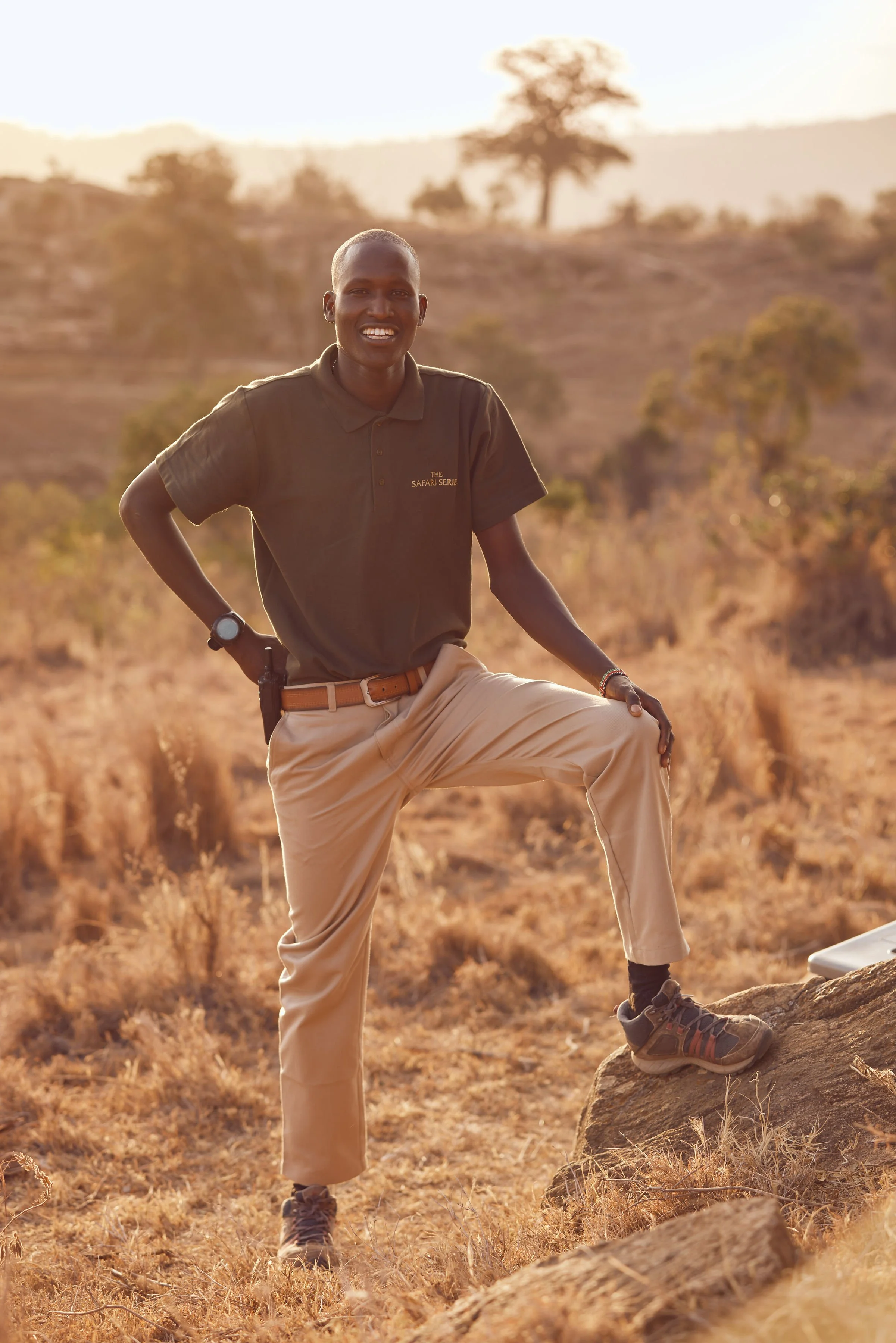 A safari guide wearing beige pants, a black T-shirt, and hiking shoes is standing outdoors in a dry, grassy landscape during sunset, with one leg raised on a rock and smiling at the camera.