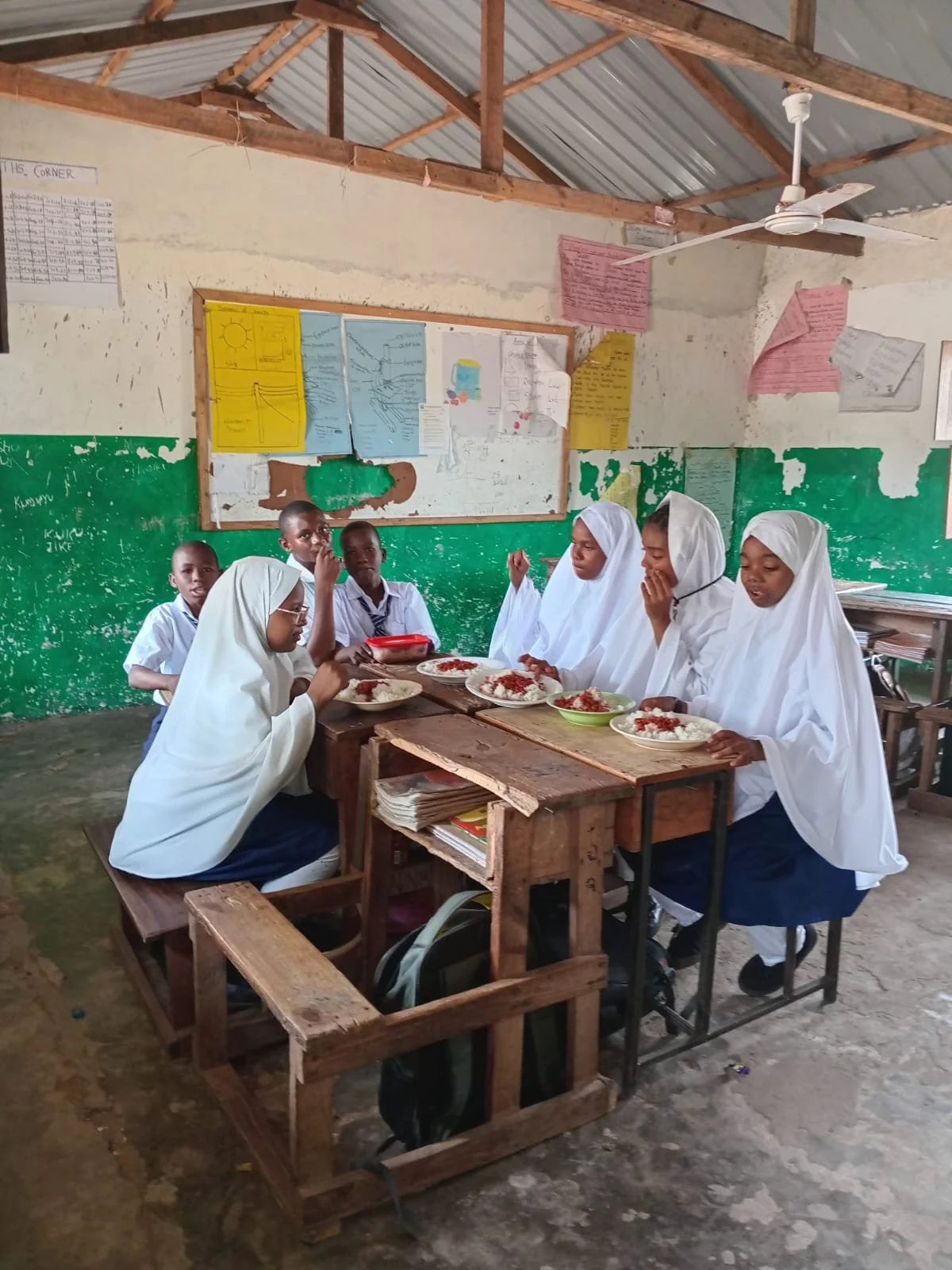School children, including girls in hijabs, sitting around a wooden desk eating a meal in a classroom with a green and white wall and educational posters.