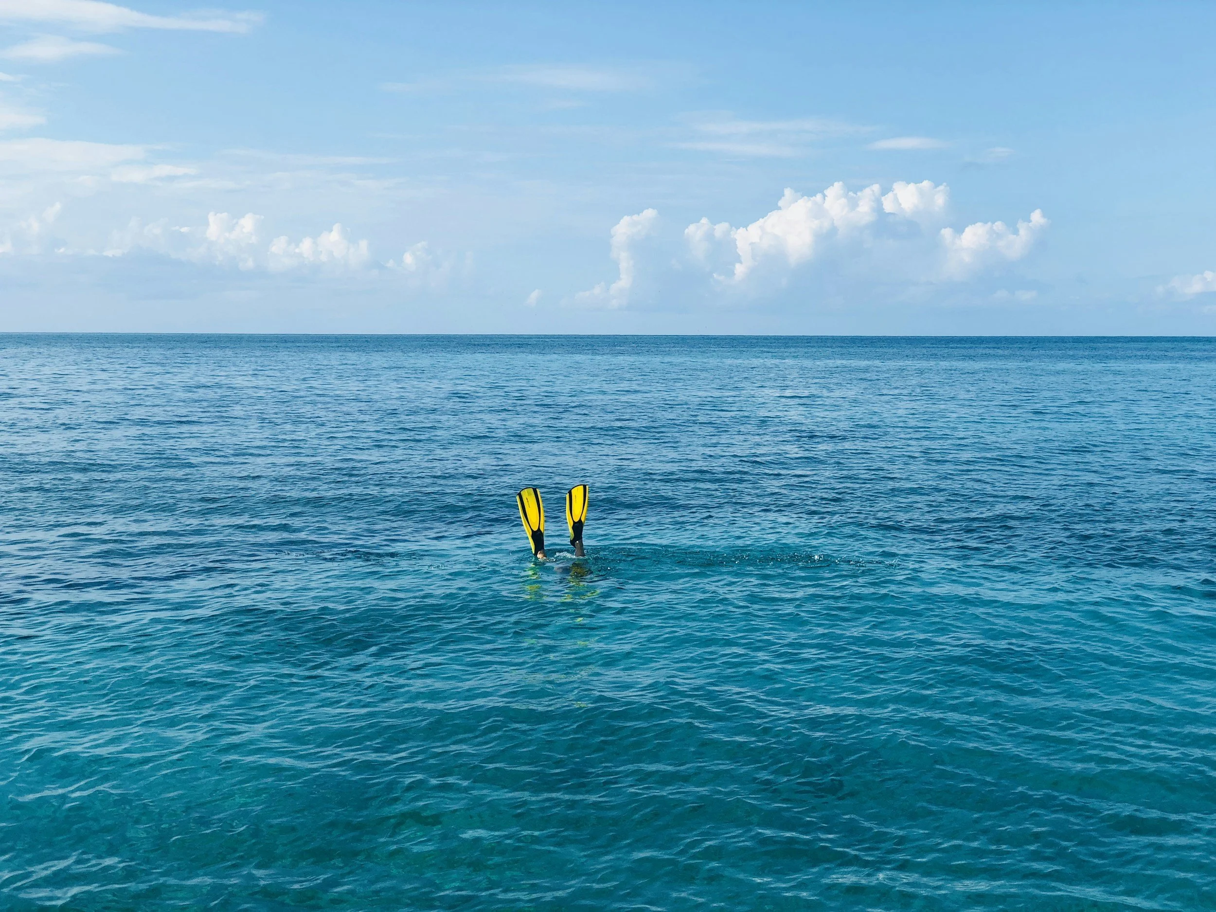 Open ocean with blue sky and white clouds, a person swimming underwater with yellow fins, visible half of their body submerged in water.