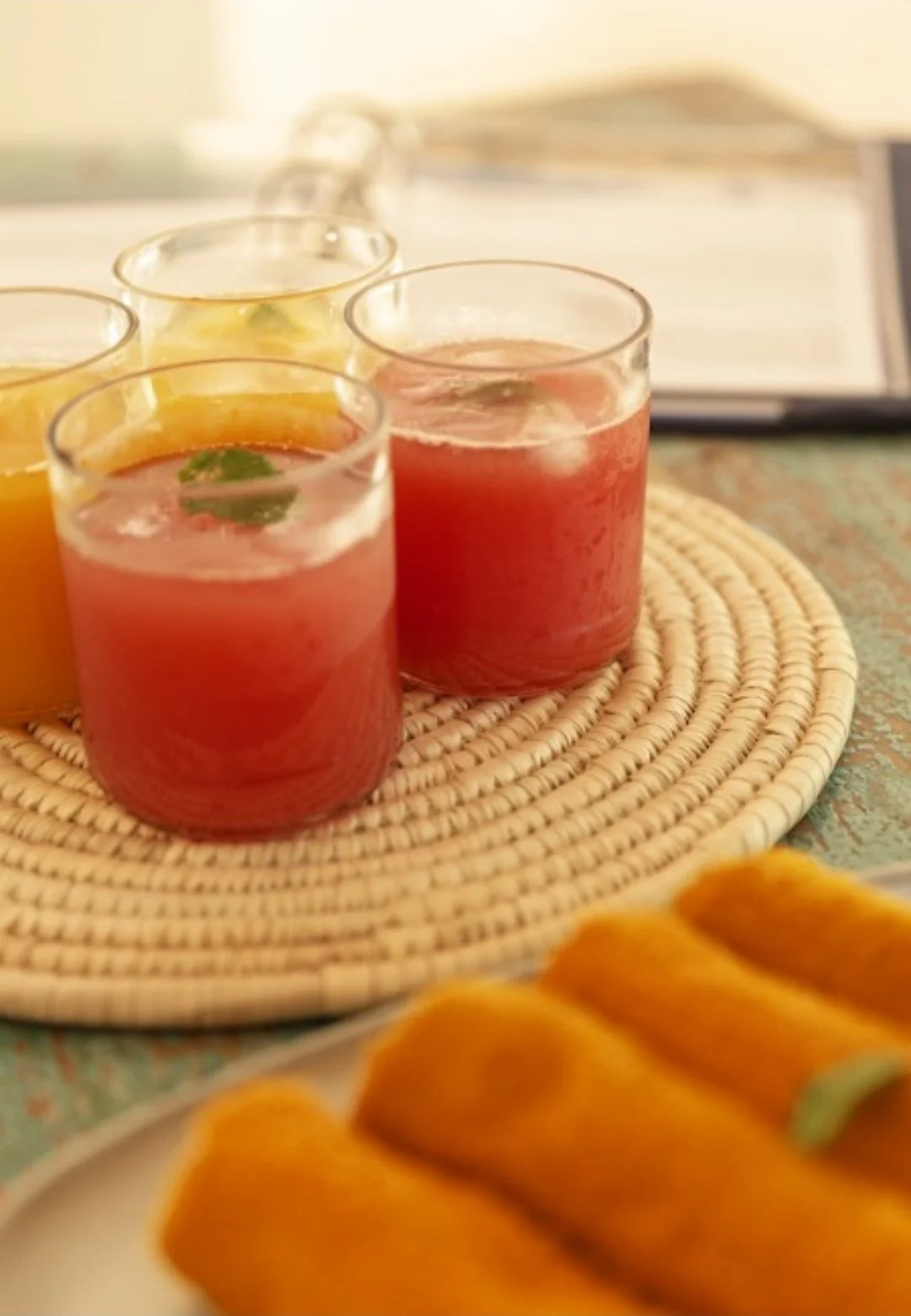 Three glasses of pink and yellow fruit drinks with mint leaves on a woven placemat, with a plate of face towels in the foreground.
