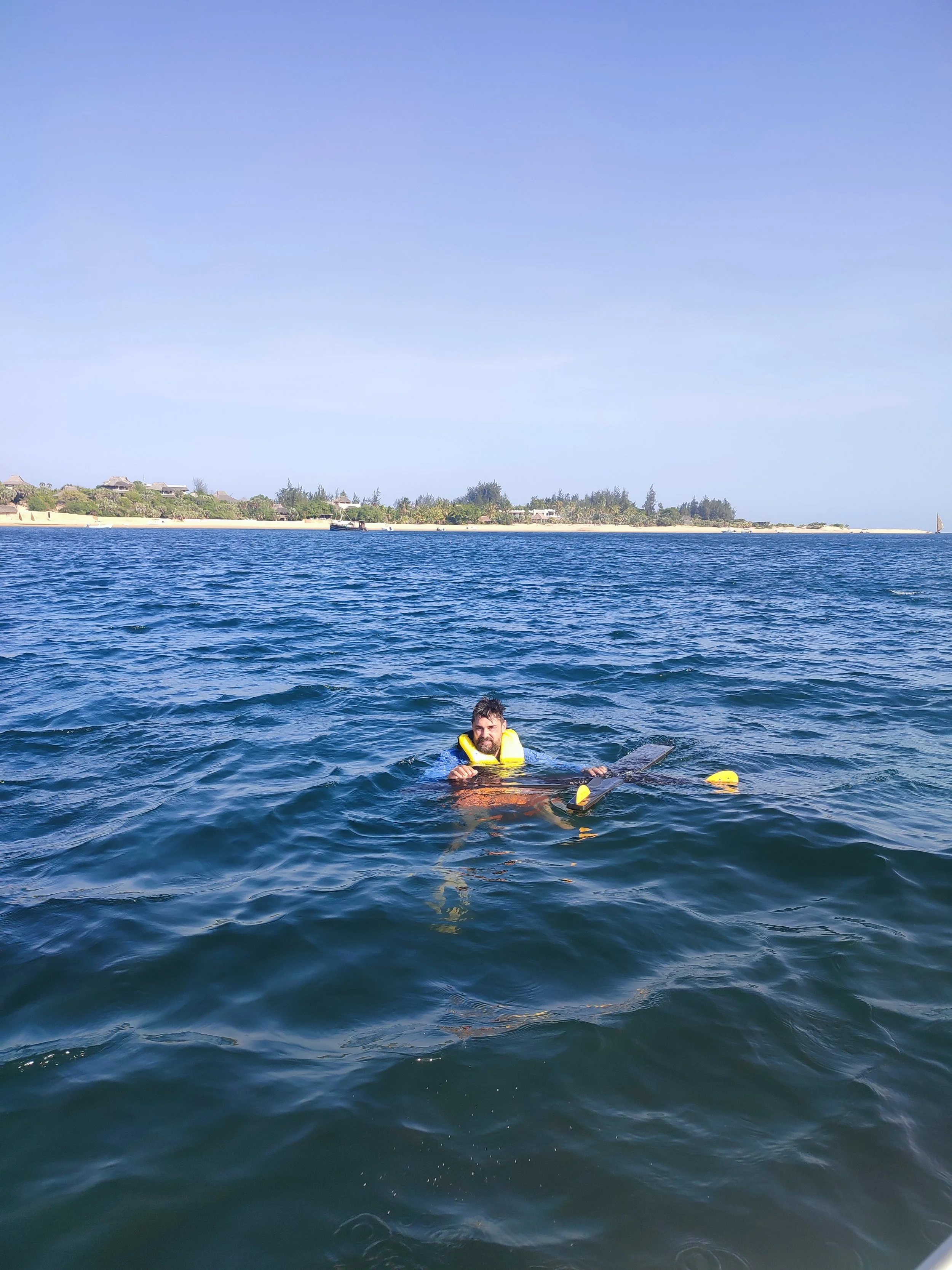 A person swimming in the ocean near the shore, wearing a yellow life jacket and holding a surfboard, with a distant shoreline and blue sky in the background.
