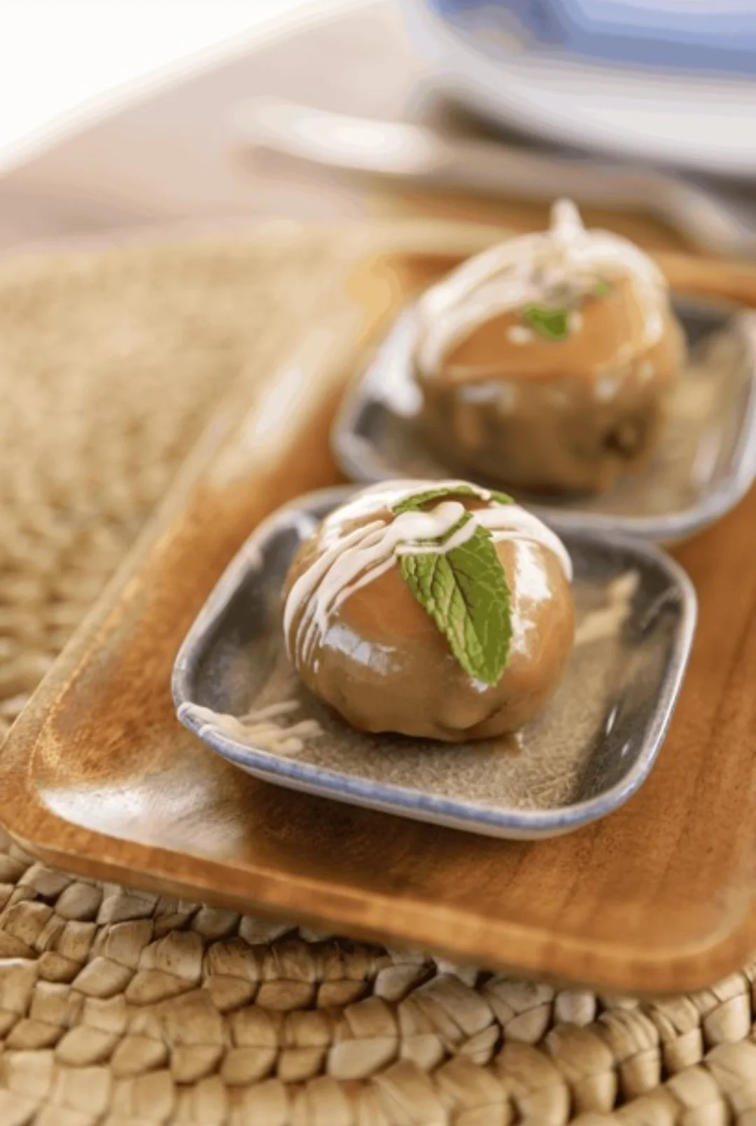 Close-up of two caramel apples topped with white drizzle and garnished with fresh mint leaves, placed on small ceramic dishes on a wooden tray.