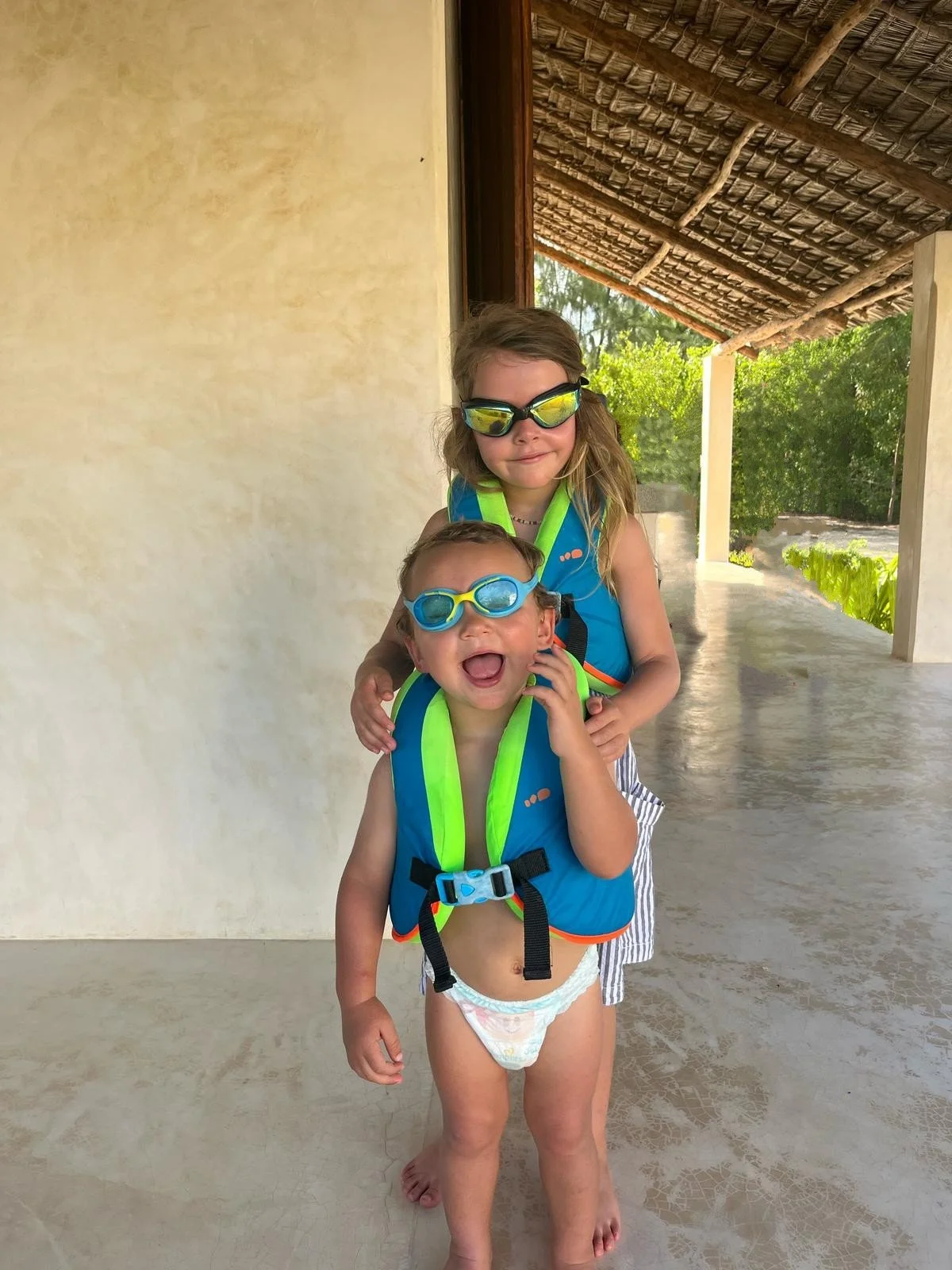 Two children, a girl and a boy, wearing life jackets and goggles, standing on a porch with a thatched roof and greenery in the background.