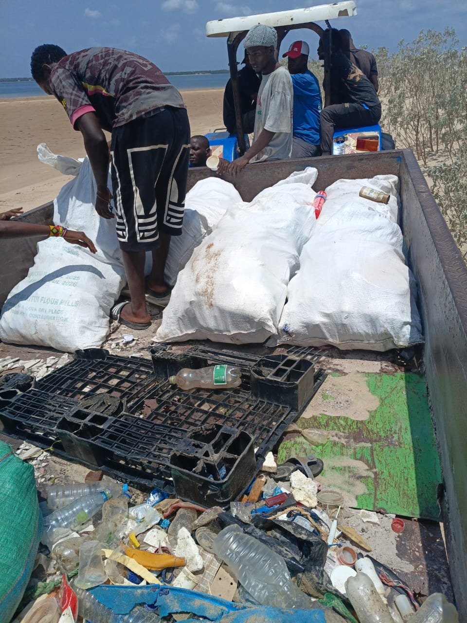 People loading large plastic bags filled with goods onto a truck on a sandy shoreline with sparse vegetation and the water in the background. The truck is surrounded by scattered trash and plastic bottles.
