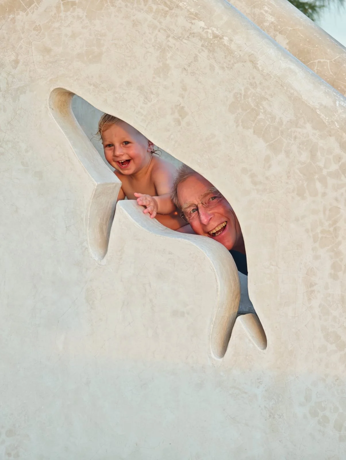 A young child and an elderly man smiling and playing behind a large decorative star-shaped cutout on a beige stone or concrete surface.