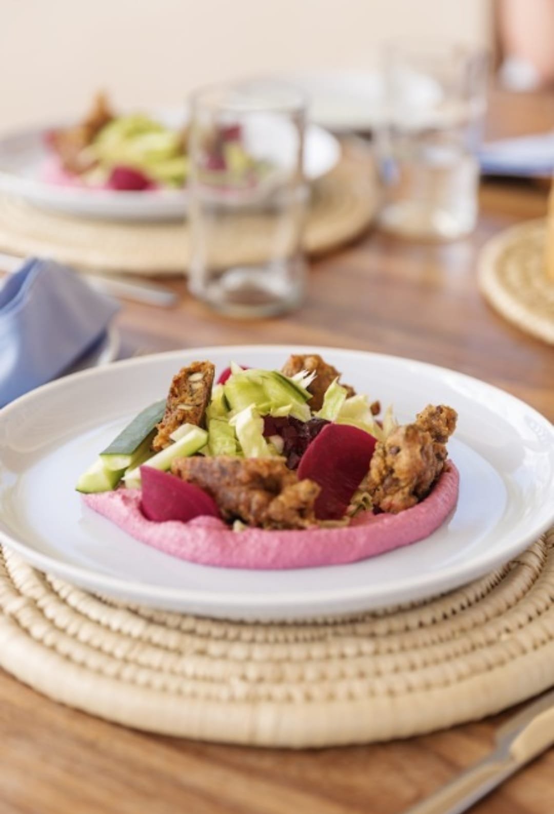 A plate with a pink tostada topped with fried chicken pieces, chopped lettuce, and radishes, on a woven placemat with a table setting in the background.