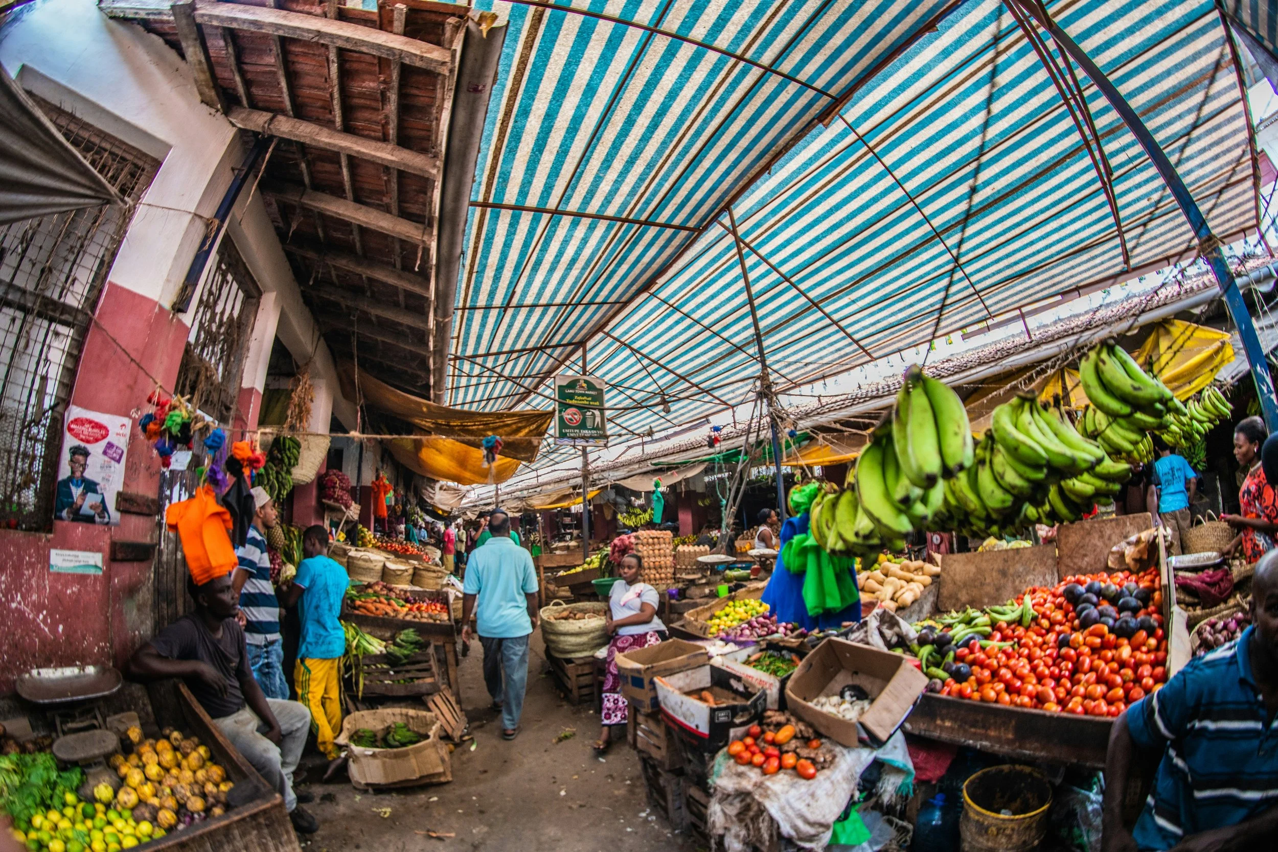 A busy outdoor market with vendors selling fresh fruits and vegetables under a striped canopy, with shoppers walking among the stalls.
