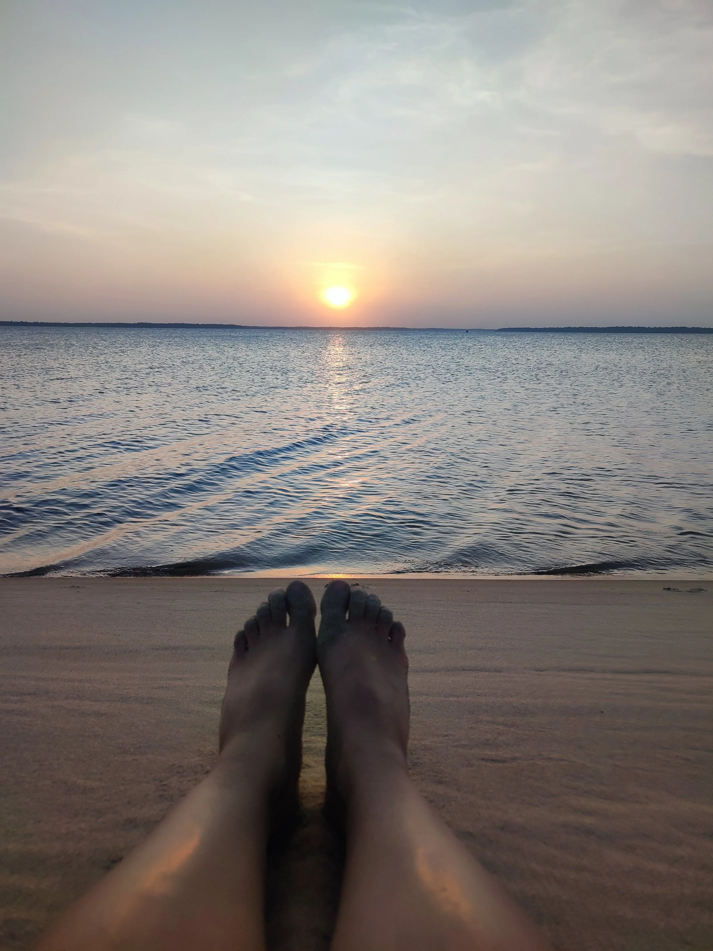 Person relaxing on a beach during sunset, with their legs stretched out on the sand and the ocean in the background.