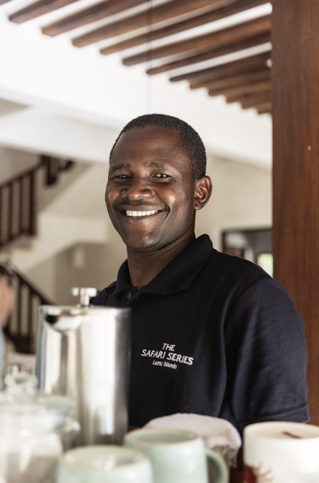 Smiling man in a black polo shirt with the text 'The Safari Series' on it, standing behind a breakfast buffet counter with tea and coffee cups.
