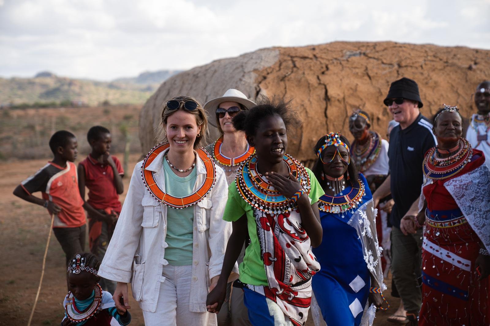 Group of women and children dressed in colorful traditional Maasai attire, walking outdoors on a sunny day with a mountain and open landscape in the background.