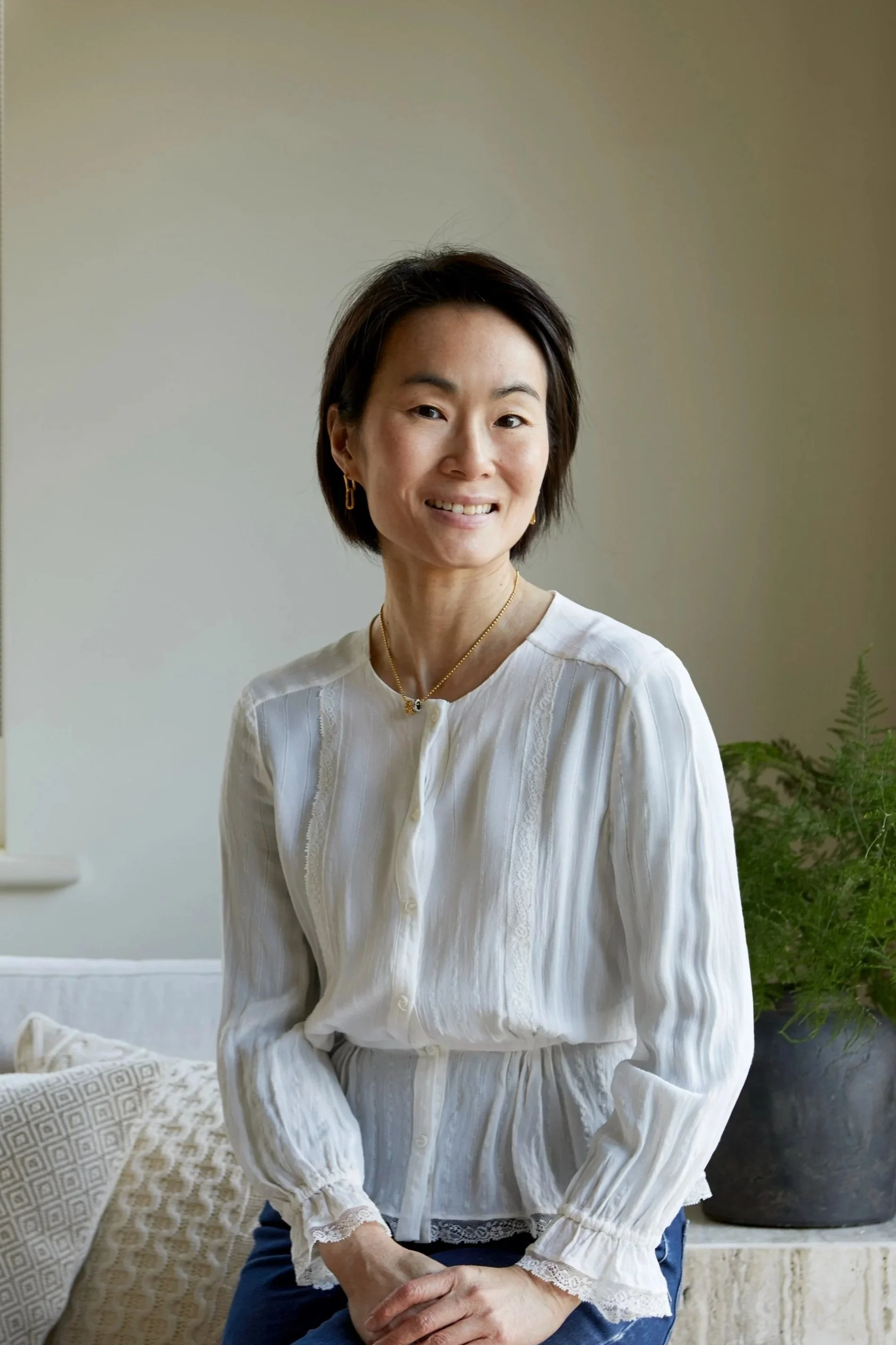 A woman with short dark hair wearing a white blouse, sitting in a room with a beige wall and some green plants in the background.