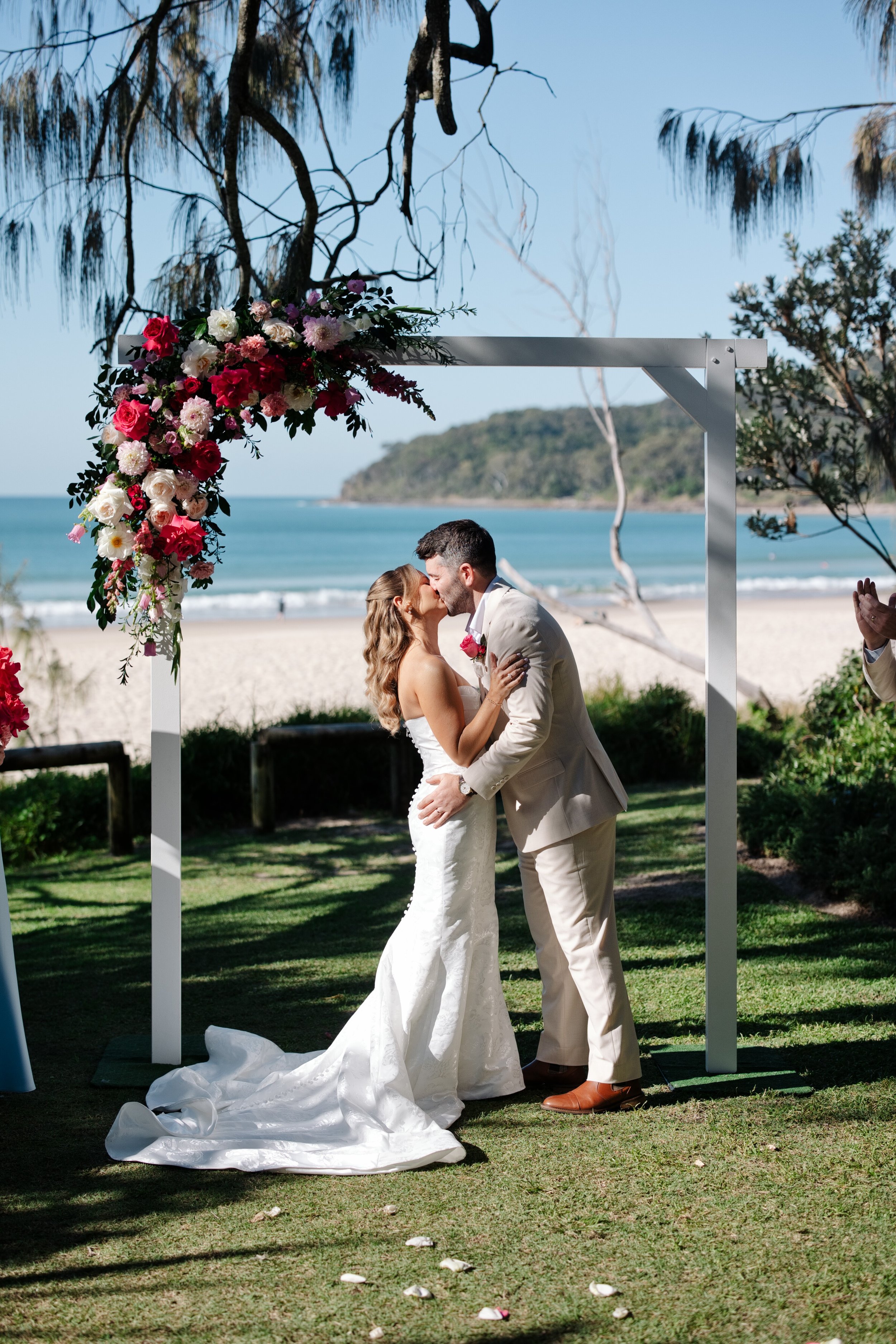 A couple dressed in wedding attire standing on a wooden walkway at the beach, with the ocean, sandy shore, and green hills in the background on a sunny day.