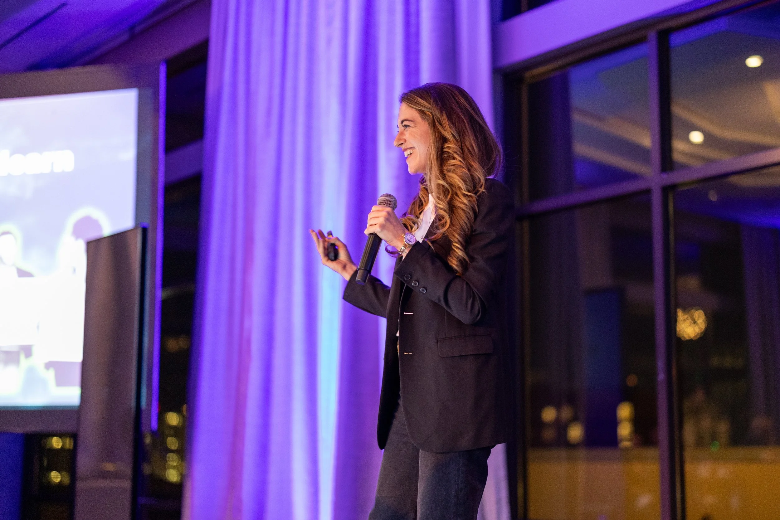 A woman with long, wavy brown hair speaking into a microphone at a conference or event, with decorative ceiling lights and an audience in the background.