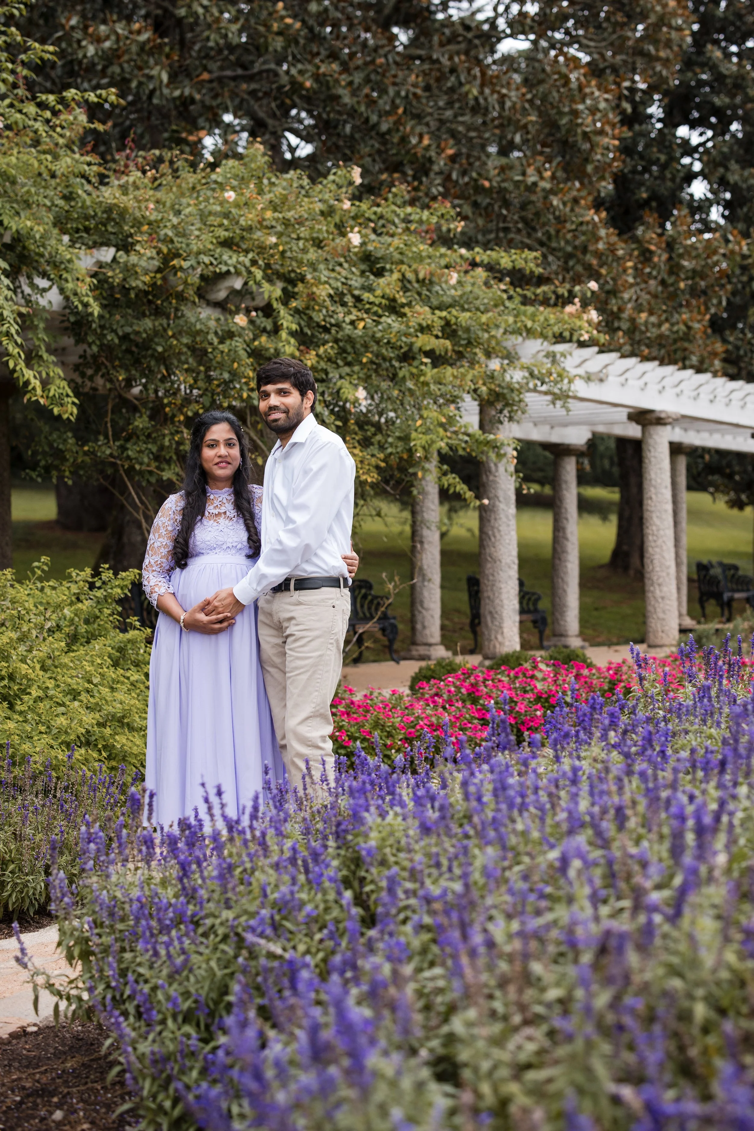 A couple standing together in a garden surrounded by purple and pink flowers, with green trees and a white pergola in the background.