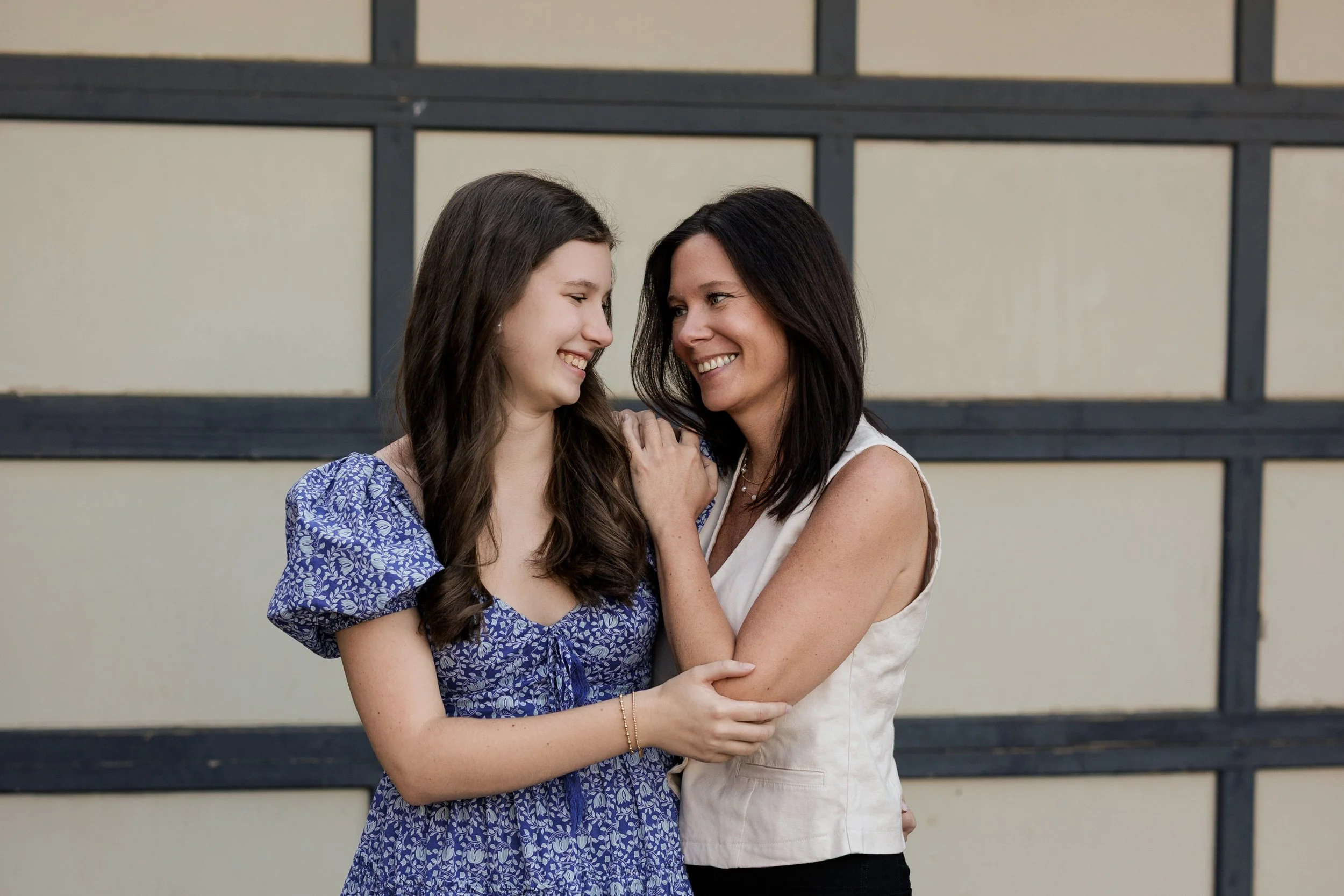 A woman and a teenage girl embracing and looking at each other joyfully, standing in front of a decorative wall with horizontal and vertical black bars.