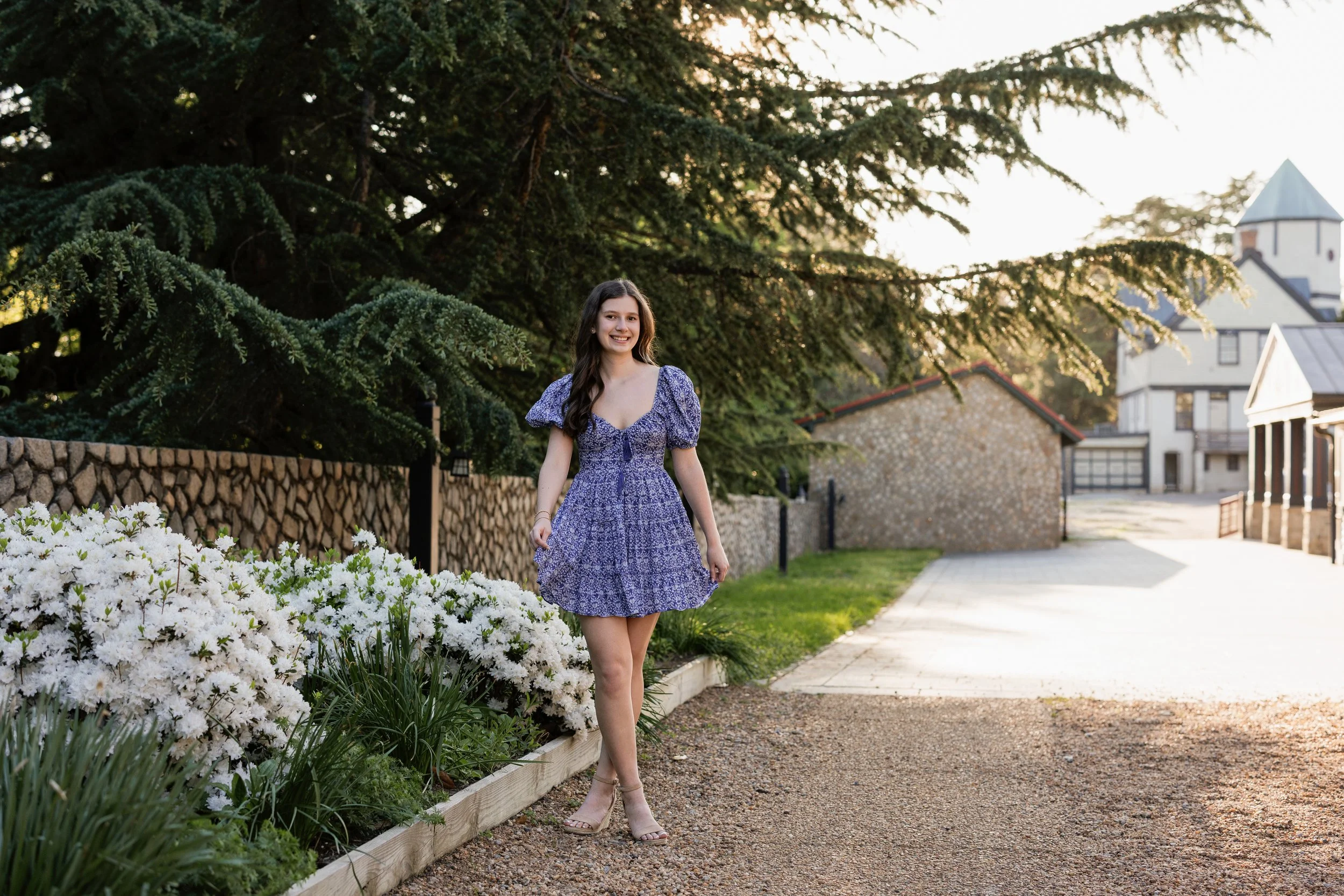 A young woman with long brown hair in a blue floral dress standing outdoors near white flowers and a large green tree, with houses and a church in the background during sunset.